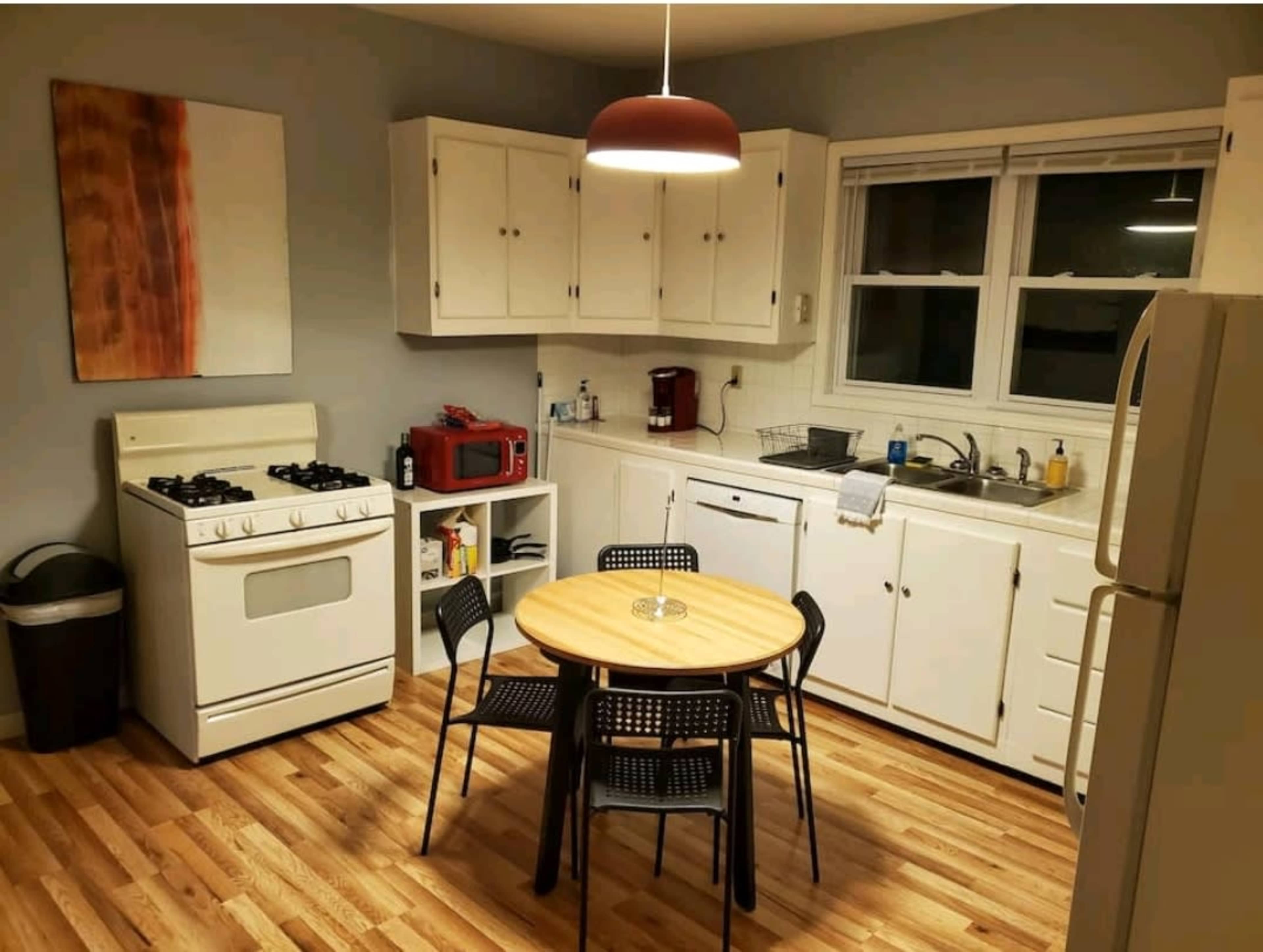 A kitchen with white cabinets, a round wooden table surrounded by four black chairs, a stove, and a sink beneath a window.