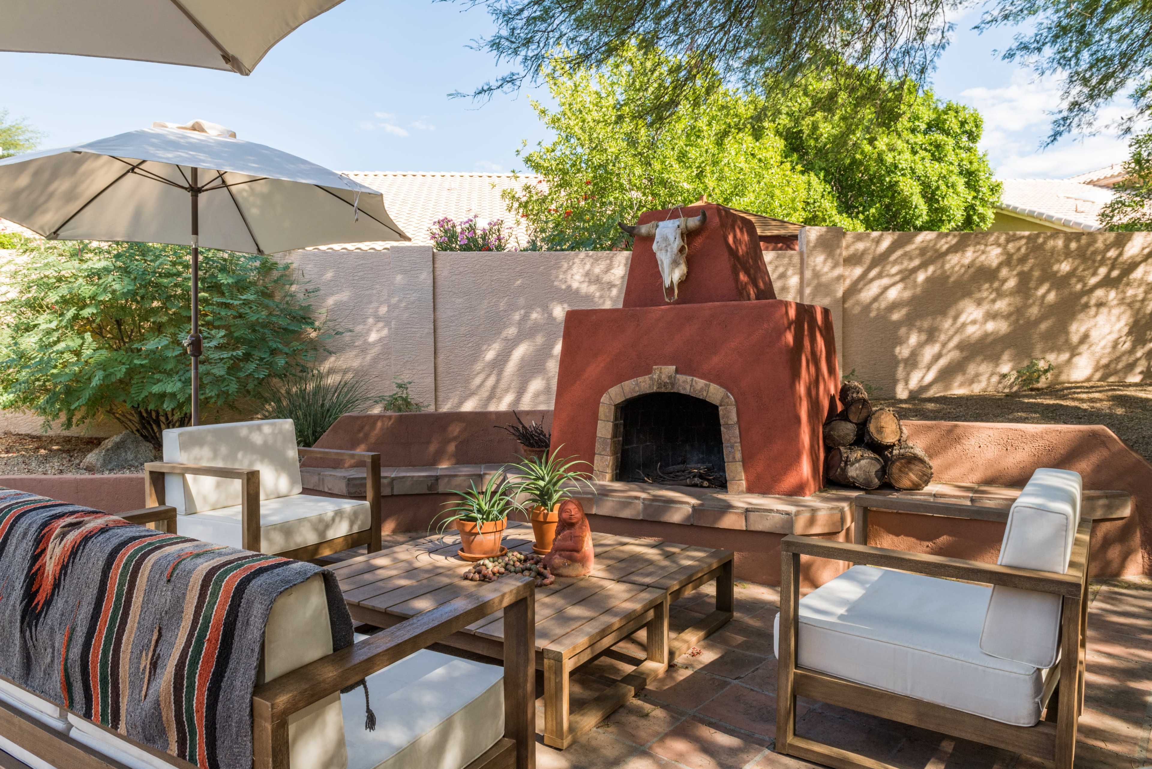 The image shows a cozy outdoor seating area with a wooden table and chairs, a fireplace adorned with a decorative bull skull, and an umbrella providing shade.