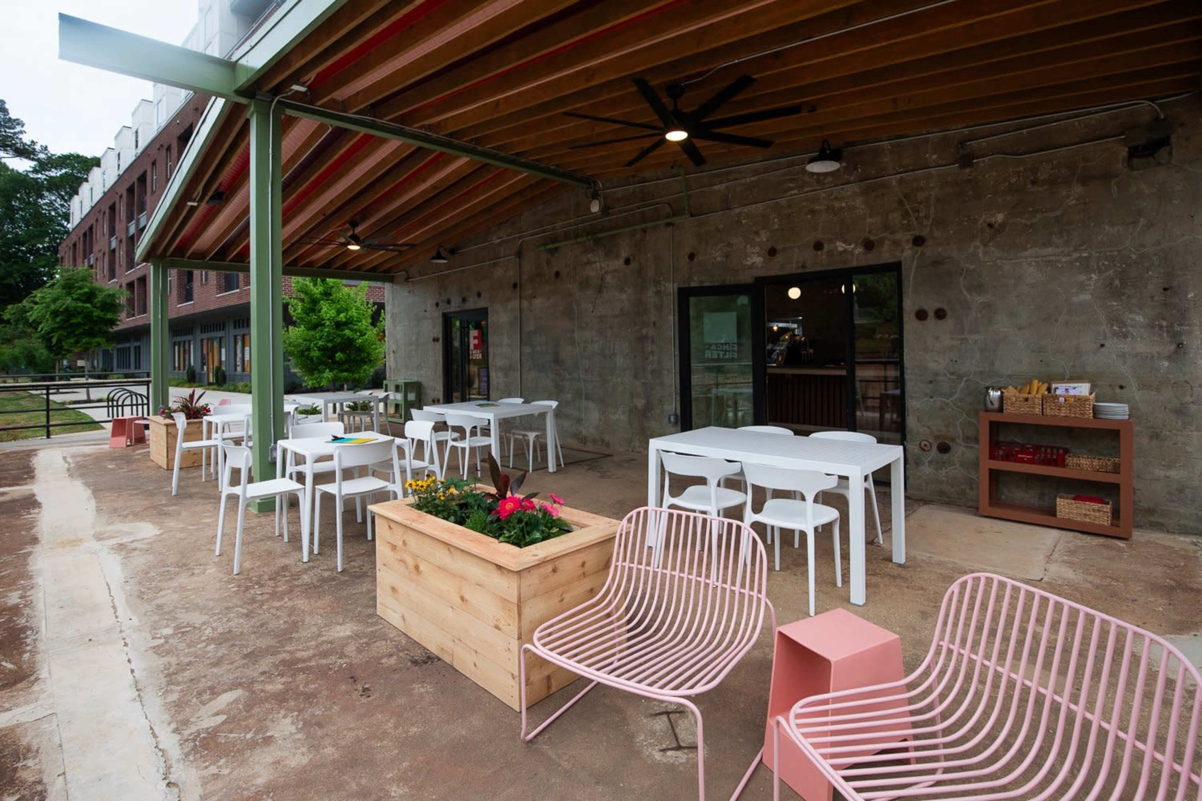 The image shows an outdoor seating area with white tables and chairs, a planter with flowers, and a concrete wall backdrop.