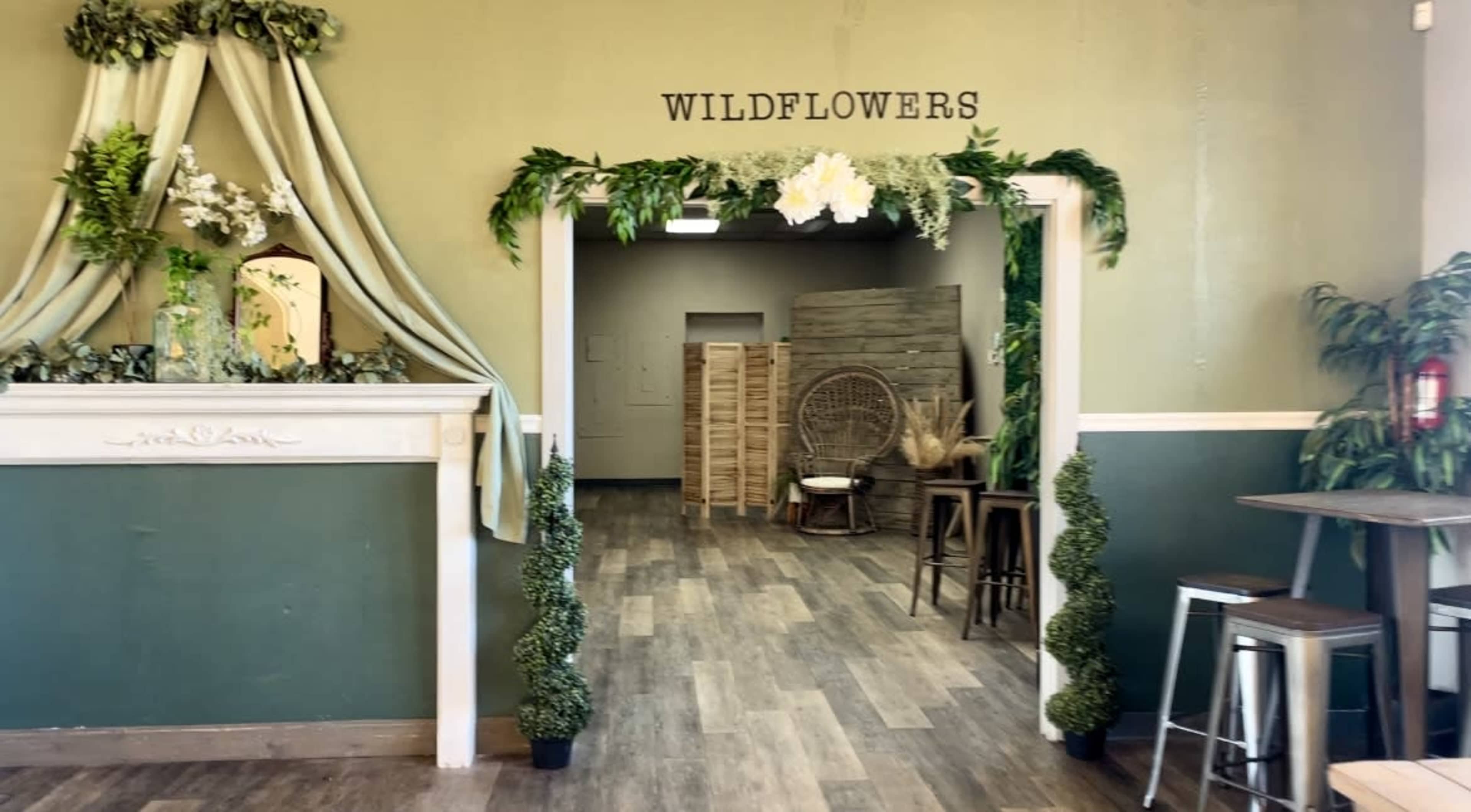 The image shows a warmly decorated indoor space with a sign that reads "WILDFLOWERS" above an entryway framed by greenery and draped fabric.
