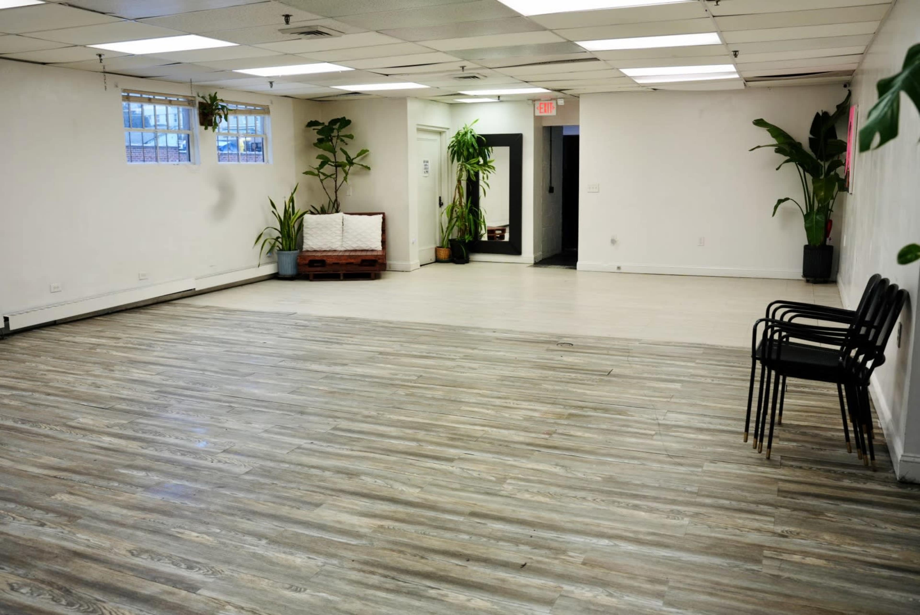 A spacious, empty room with a wooden floor, potted plants, a bench, and several black chairs stacked against the wall.