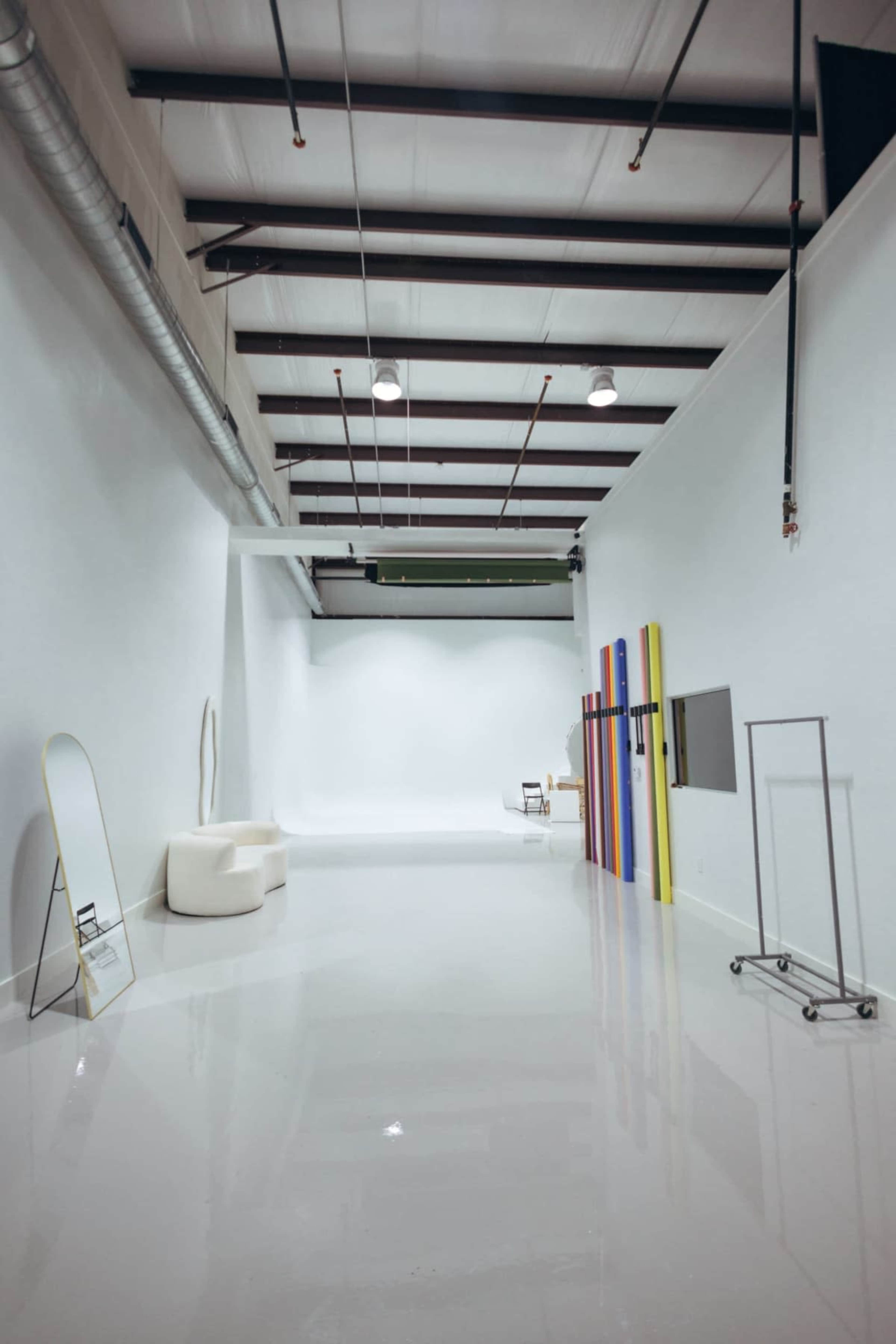 The image shows a spacious, brightly lit studio with a white background, featuring colorful backdrop rolls and minimalist furniture.