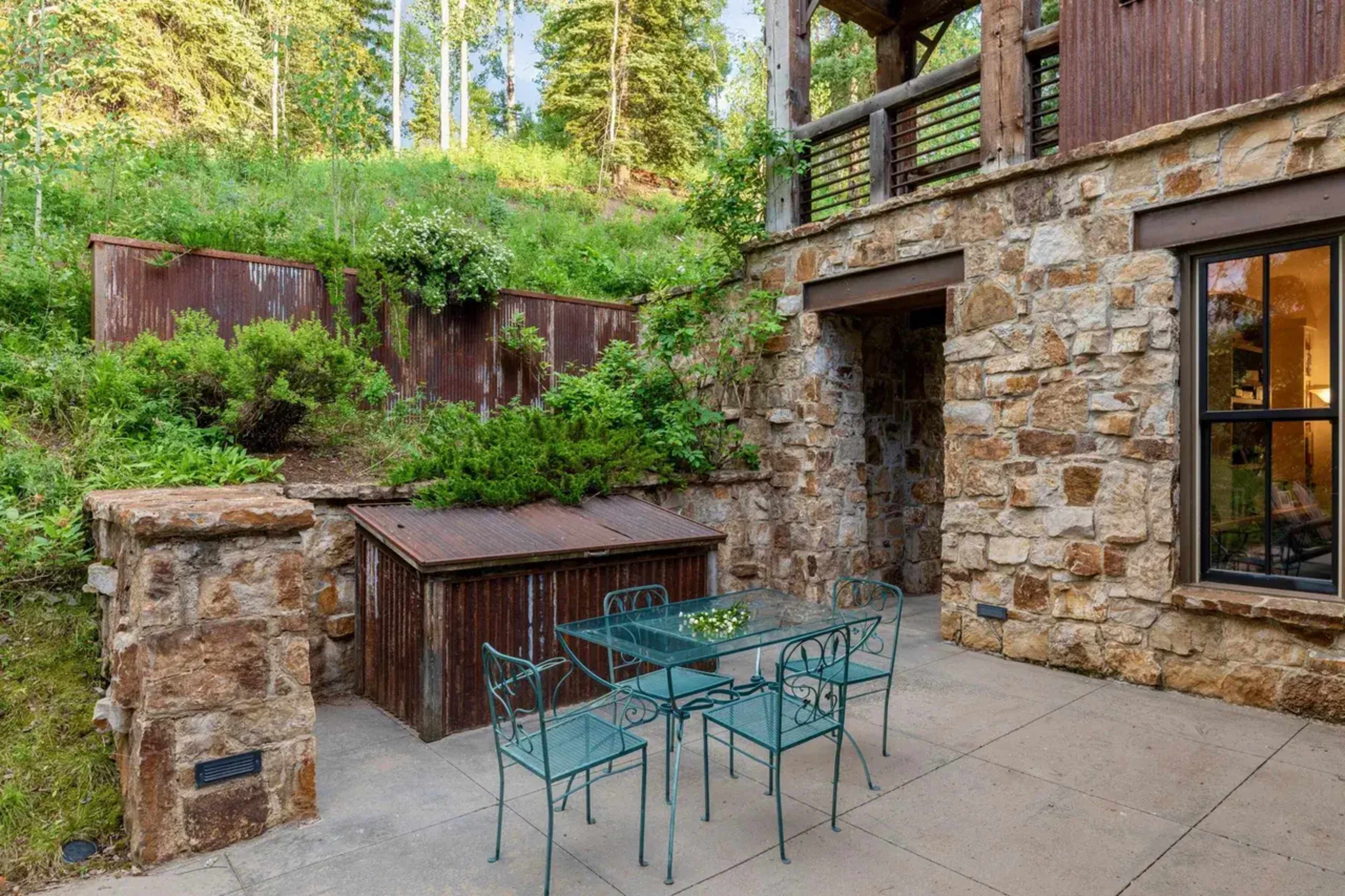 The image shows a stone patio with a metal table and chairs set against a backdrop of greenery and a rustic stone wall.