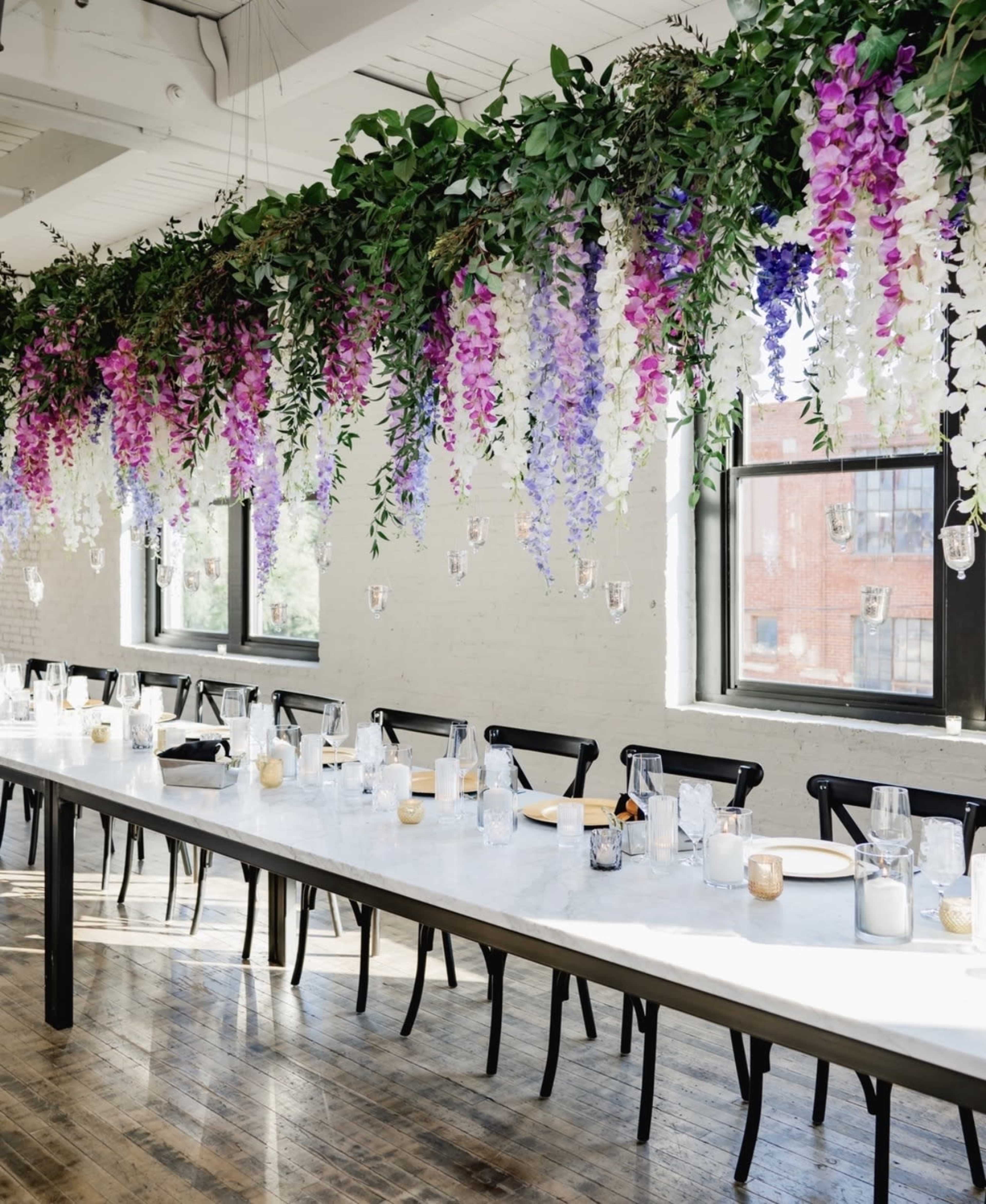 A long table is set with glassware and candles beneath a decorative overhead arrangement of flowers and greenery in a well-lit room.