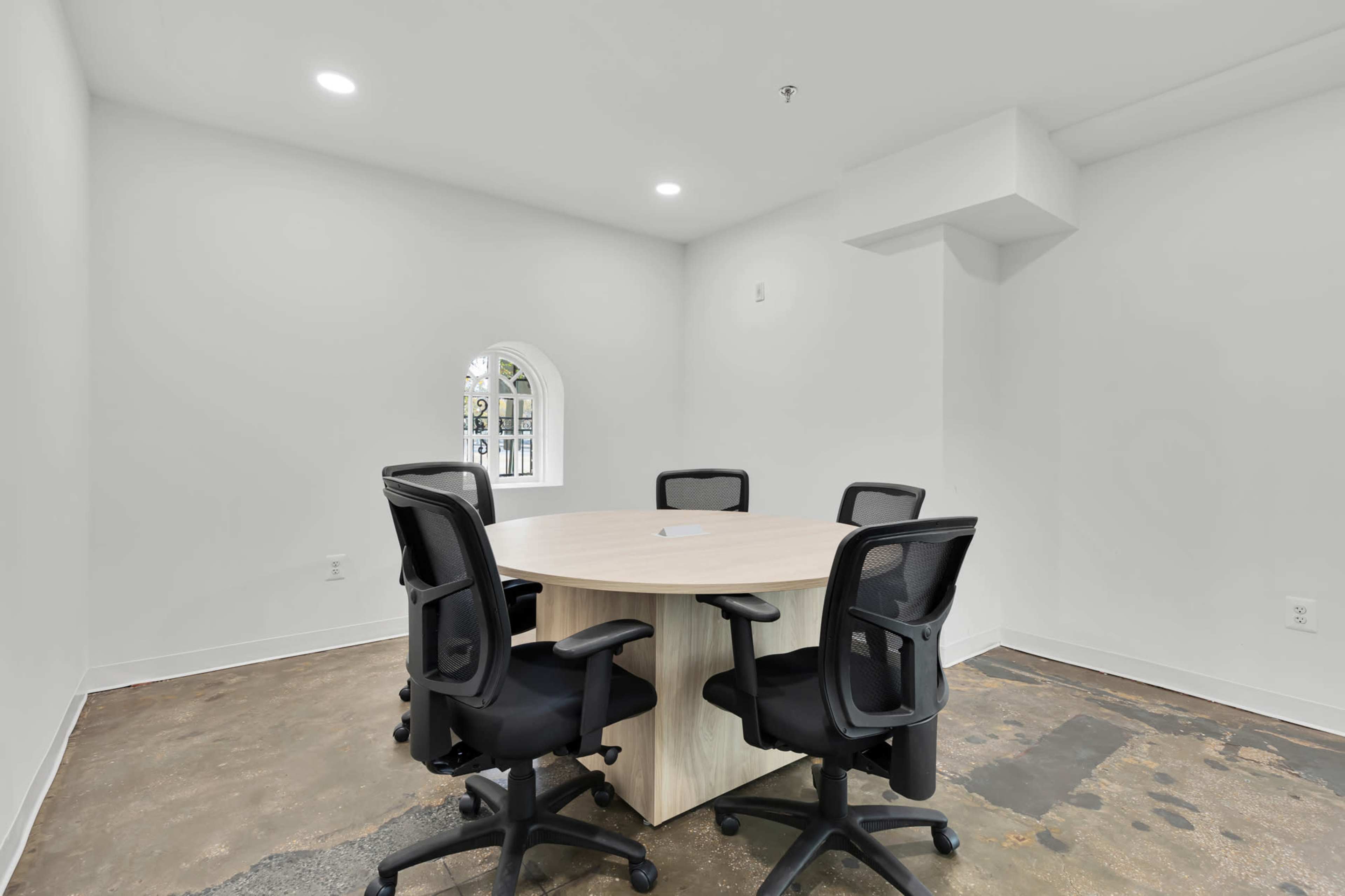 A small meeting room features a round table with five black office chairs in a bright, minimalist space.