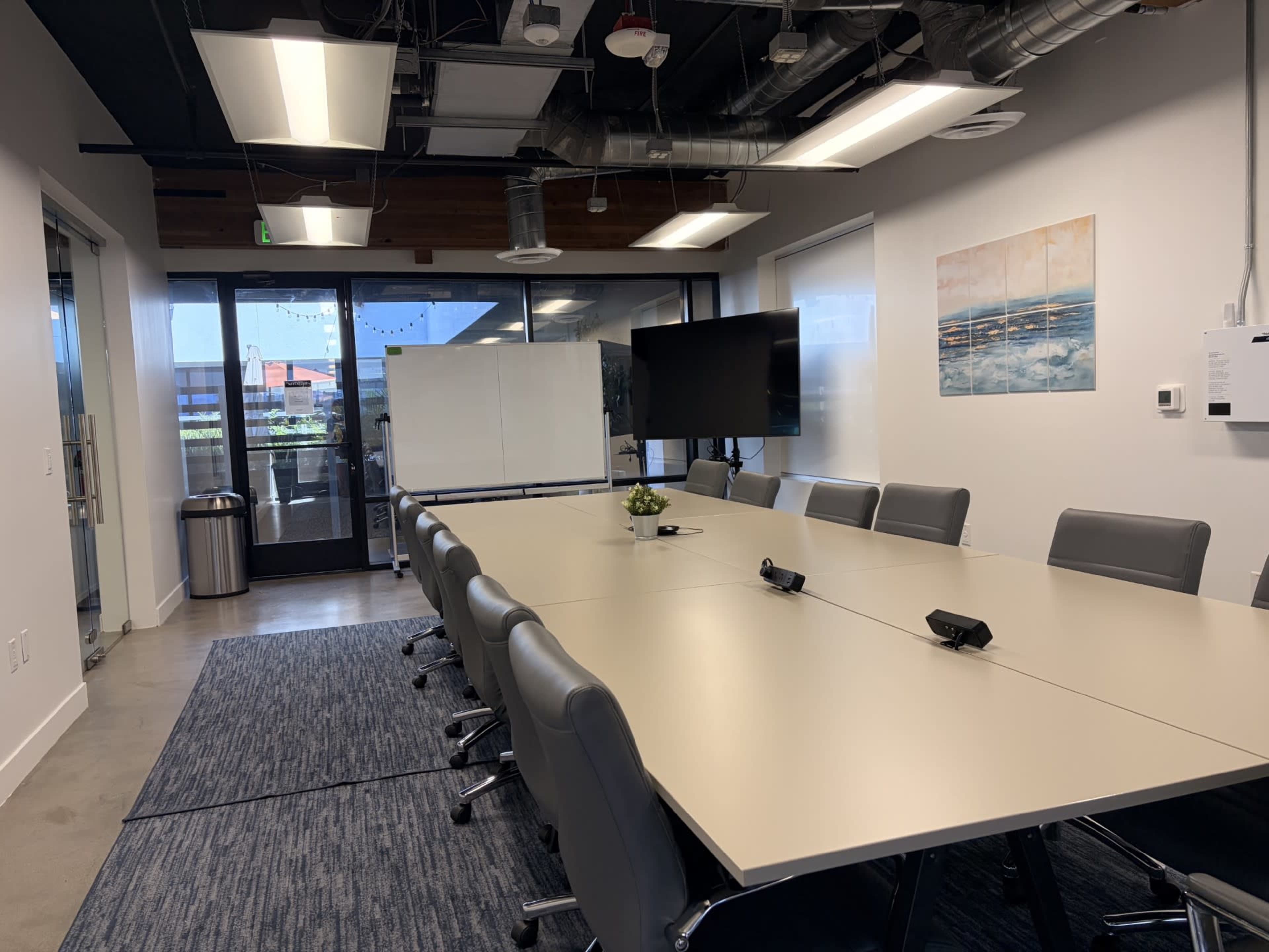 The image shows a modern conference room with a long table, ergonomic chairs, a large screen, and windows that allow natural light.