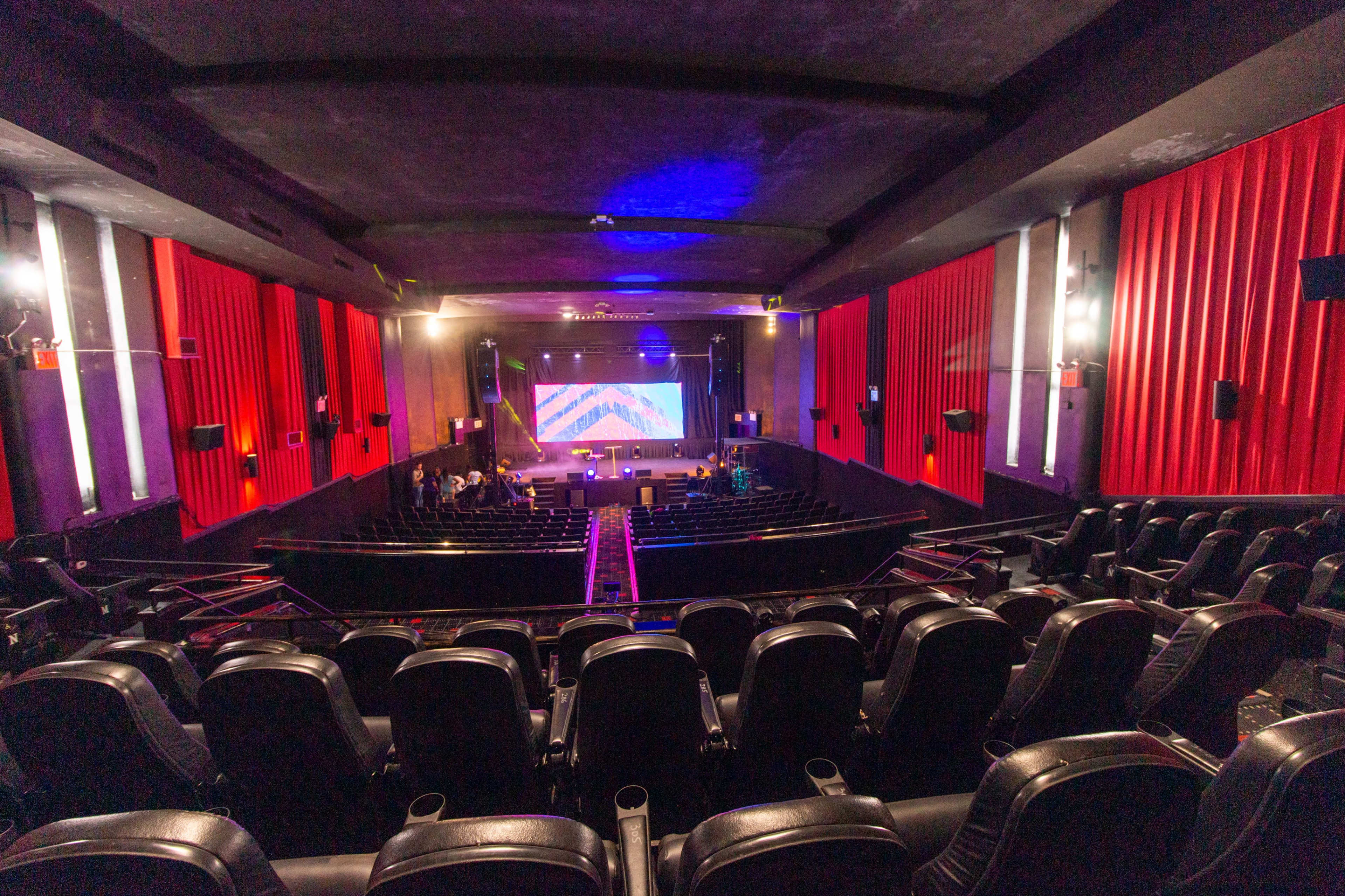 An empty theater with rows of black seating, red curtains, and a large screen illuminated in the front.