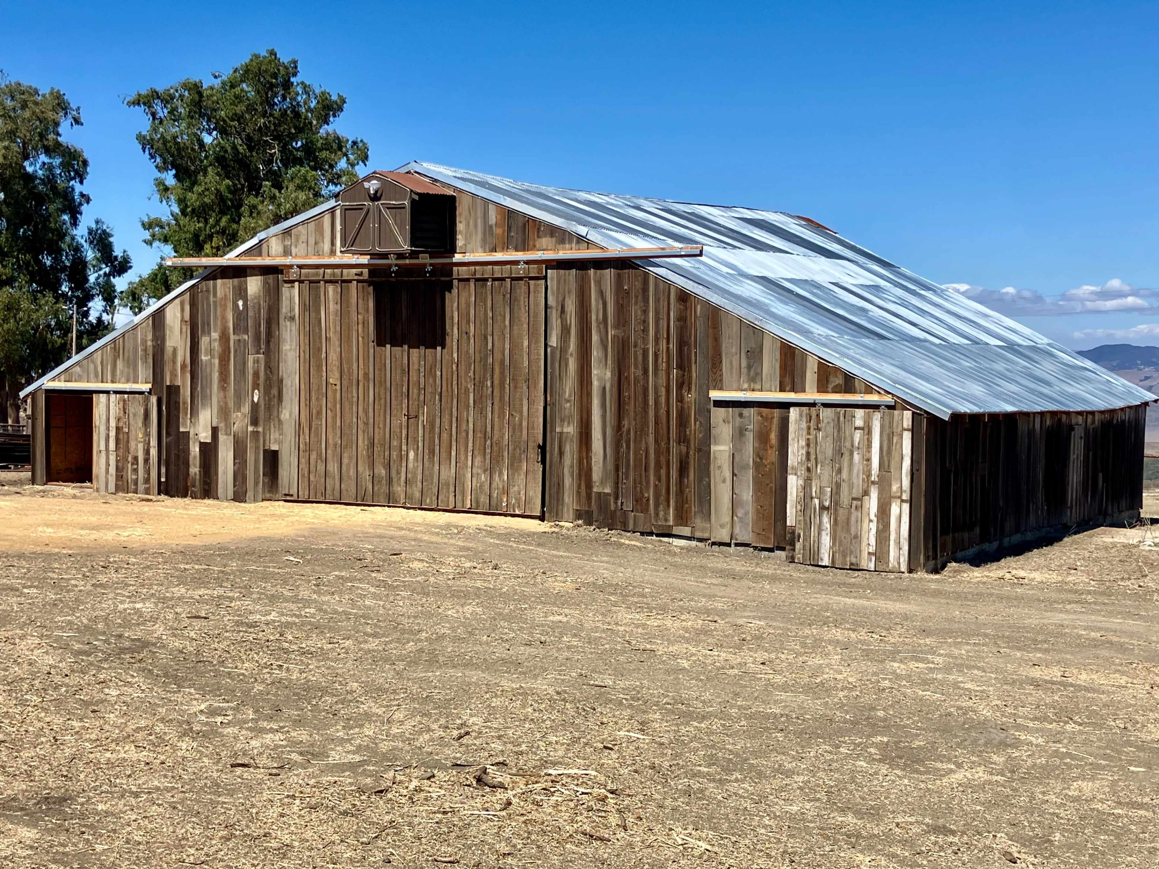 A large wooden barn with a metal roof stands in an open field under a clear blue sky.