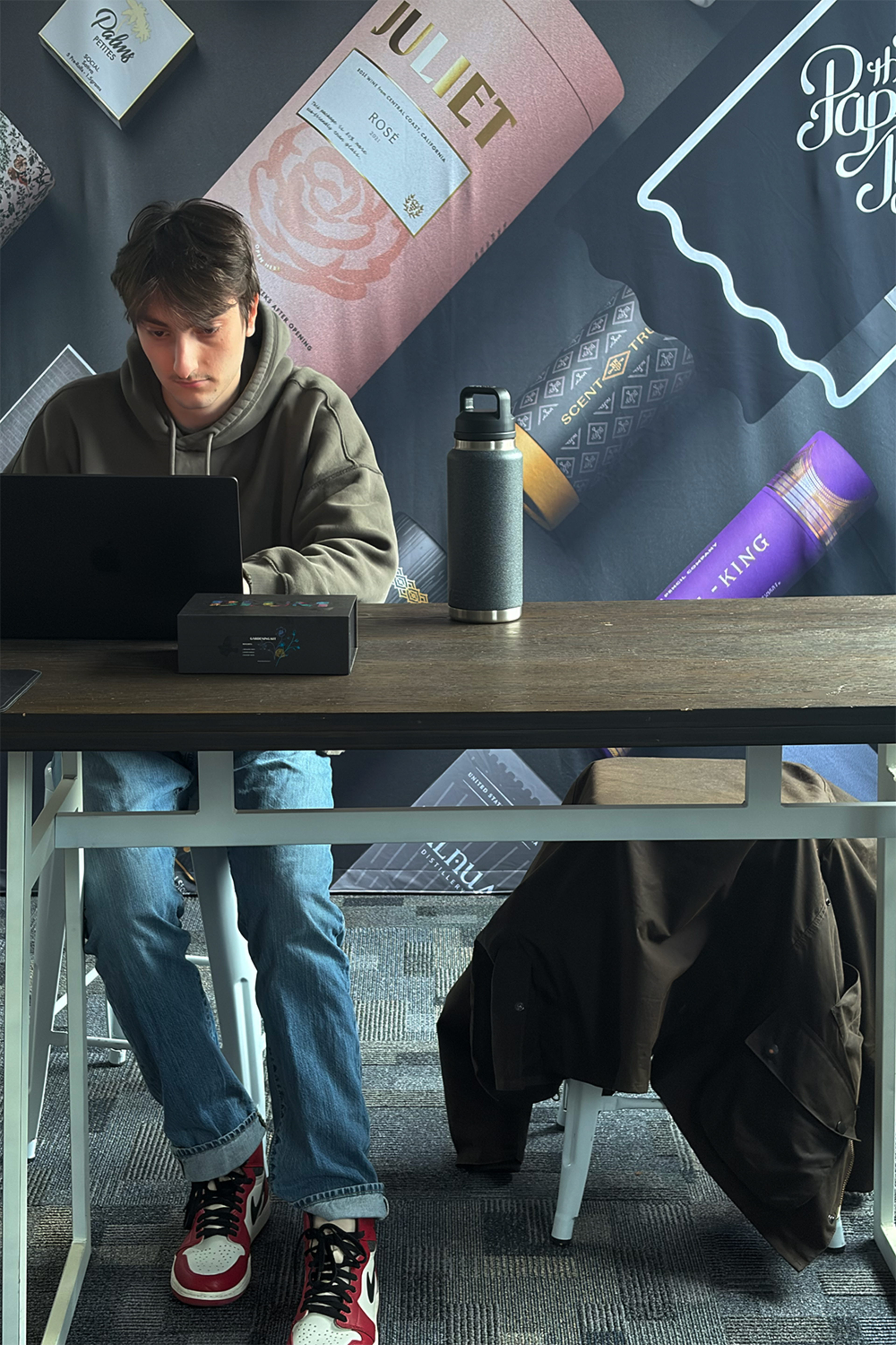 A young man sits at a wooden desk with a laptop and a water bottle, in front of a colorful backdrop featuring various product packaging.