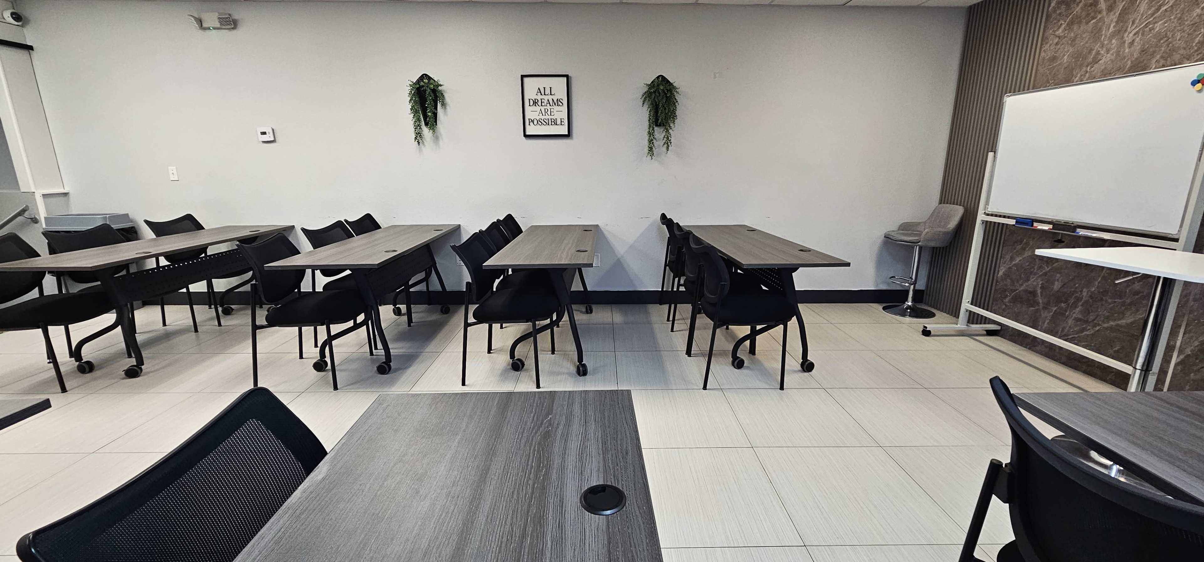 A classroom or meeting space features multiple rows of tables and chairs, with a whiteboard and decorative wall plants in the background.