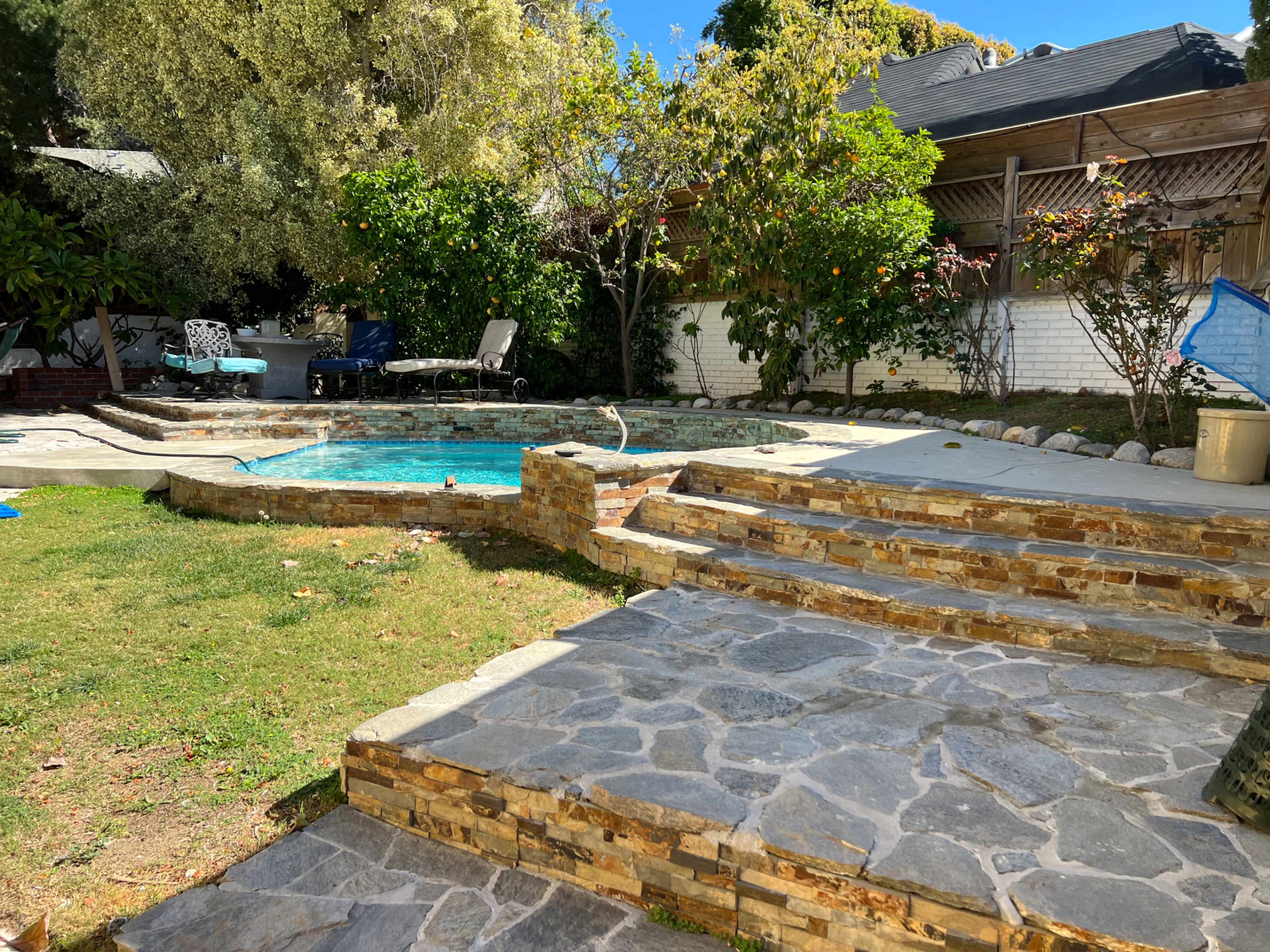 A backyard with a stone-paved area leading to a blue pool surrounded by greenery and seating.