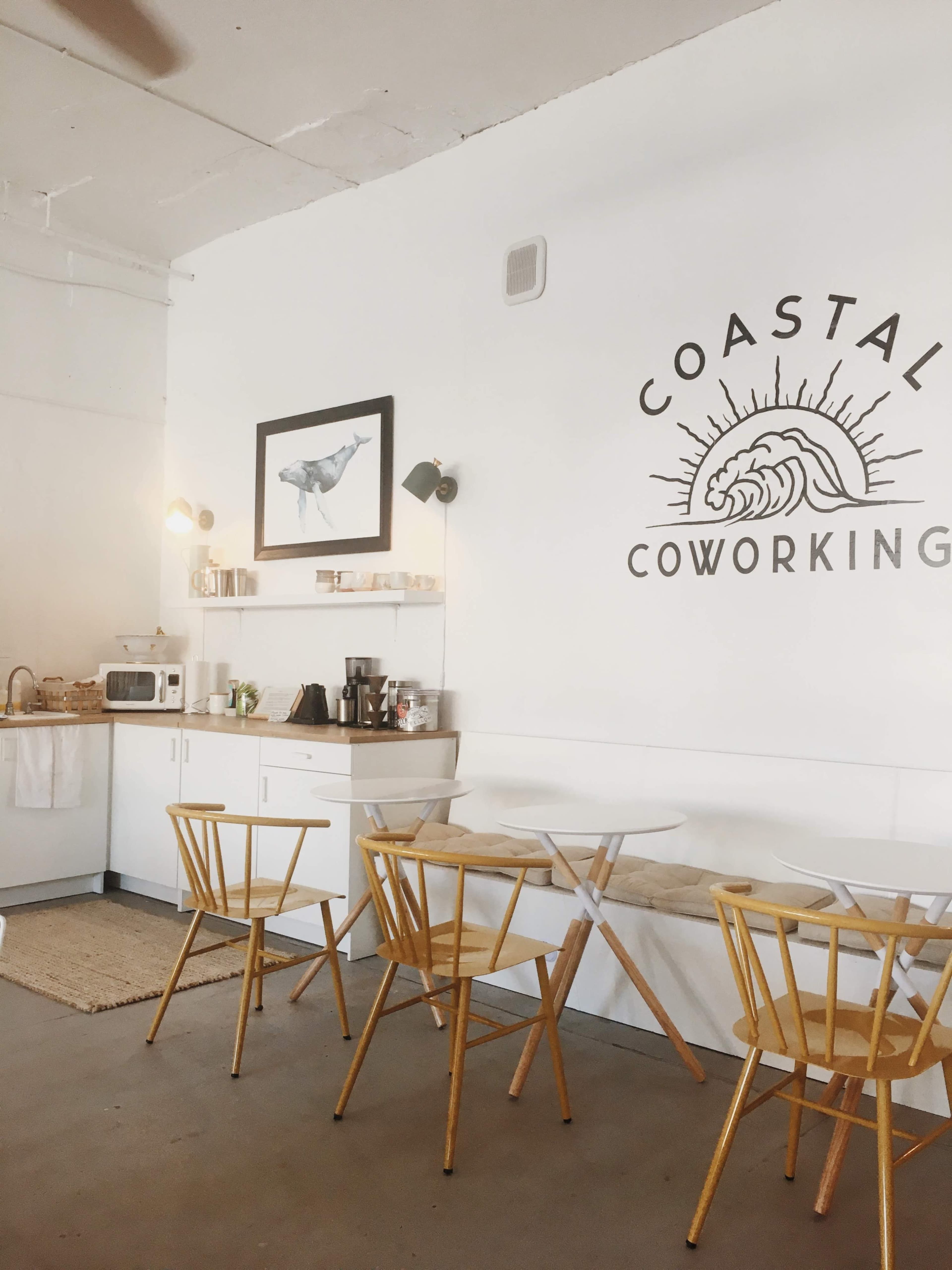 A bright, minimalist coworking space featuring a kitchen area with a microwave, a coffee station, and wooden chairs around a table against a white wall with a framed artwork and the words "COASTAL COWORKING."