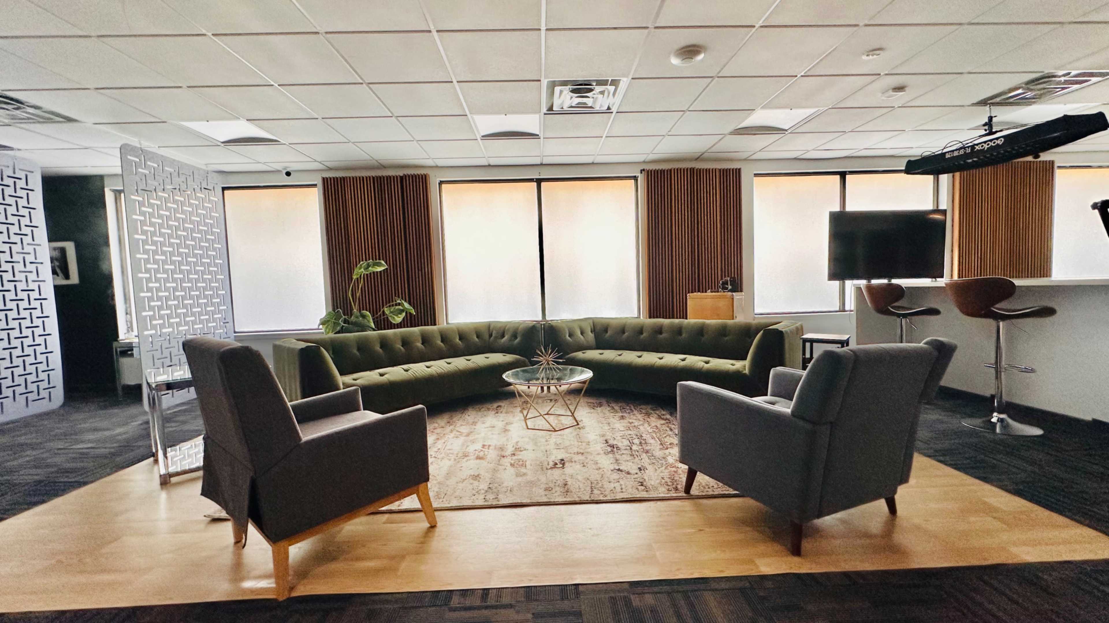 The image shows a modern office lounge area with a curved green sofa, two gray armchairs, a round coffee table, and large windows with sheer curtains.