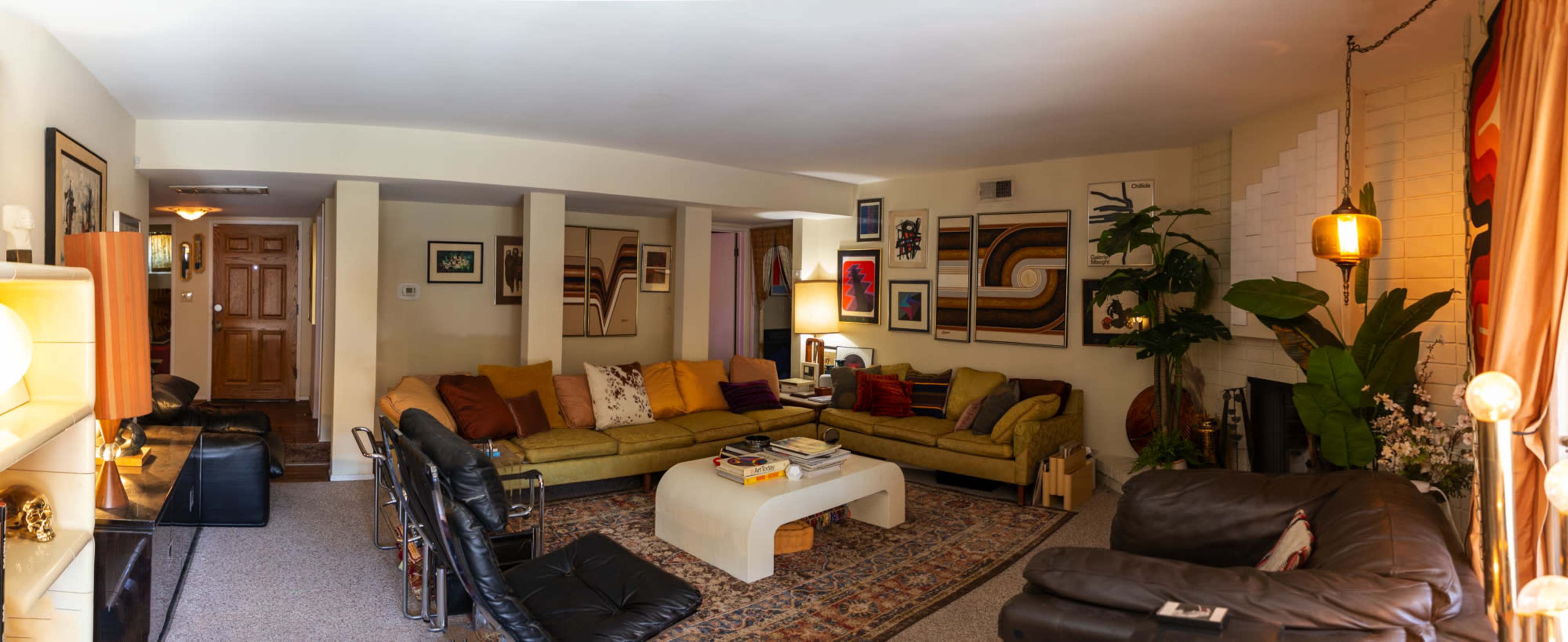 The image shows a spacious living room featuring two distinct seating areas, a coffee table, various artworks on the walls, and indoor plants.