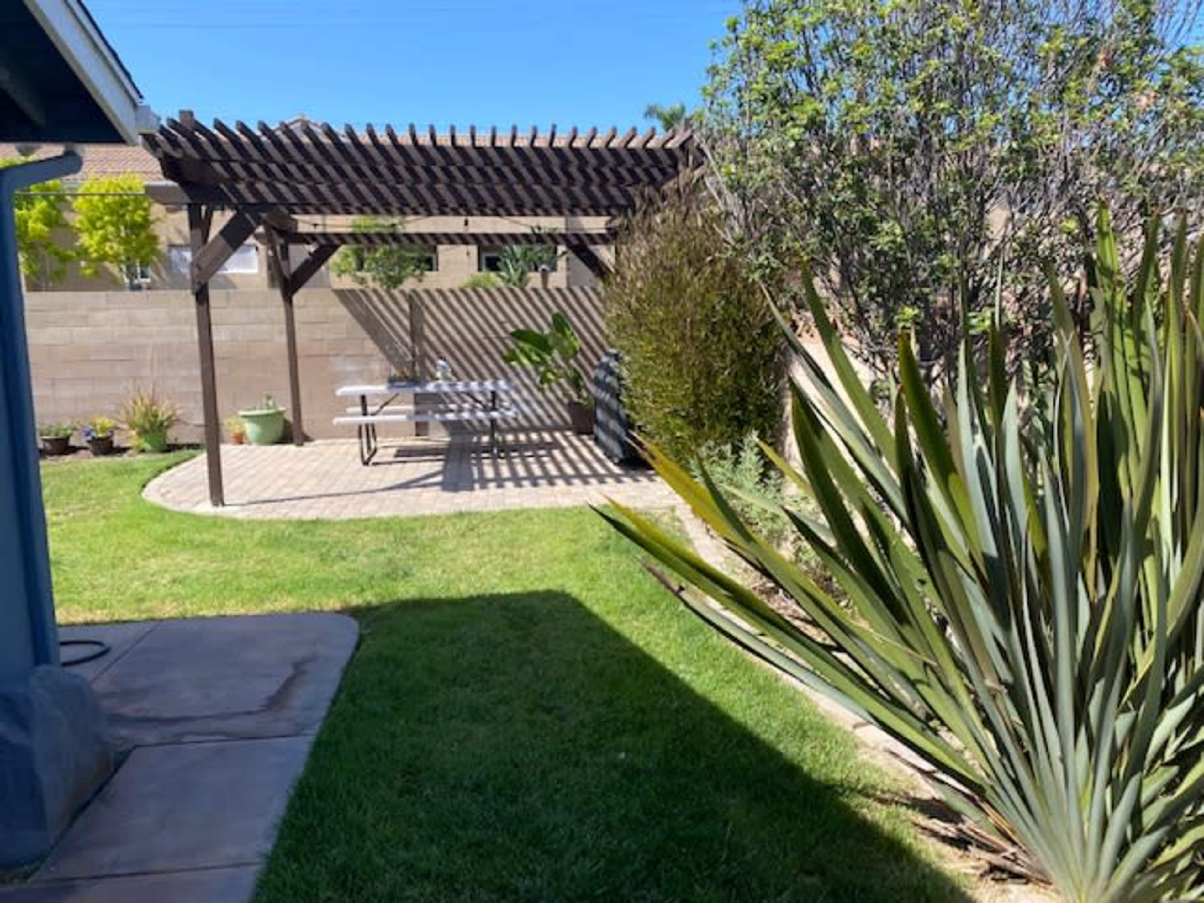 A backyard with a pergola, a stone patio with a picnic table, and landscaped greenery including large plants and grass.