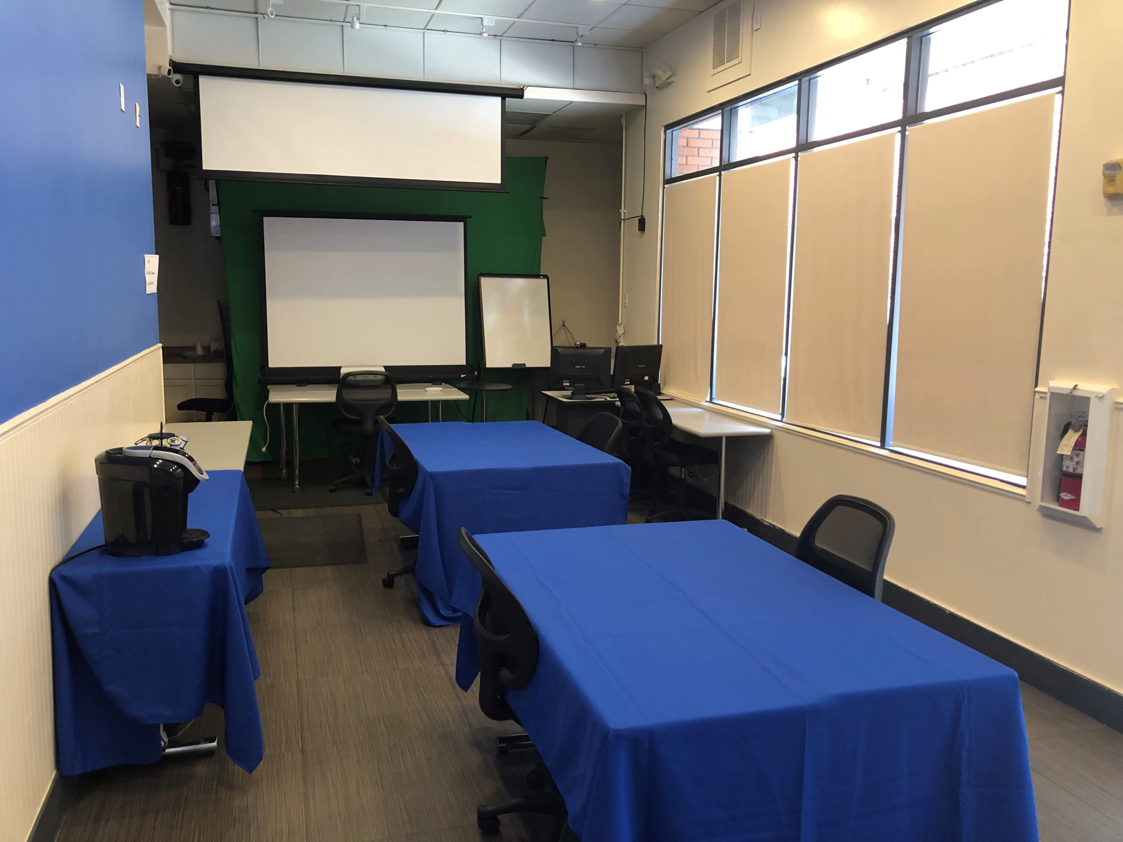 A room set up for a meeting, featuring several tables covered with blue tablecloths, a projection screen, and a green backdrop.