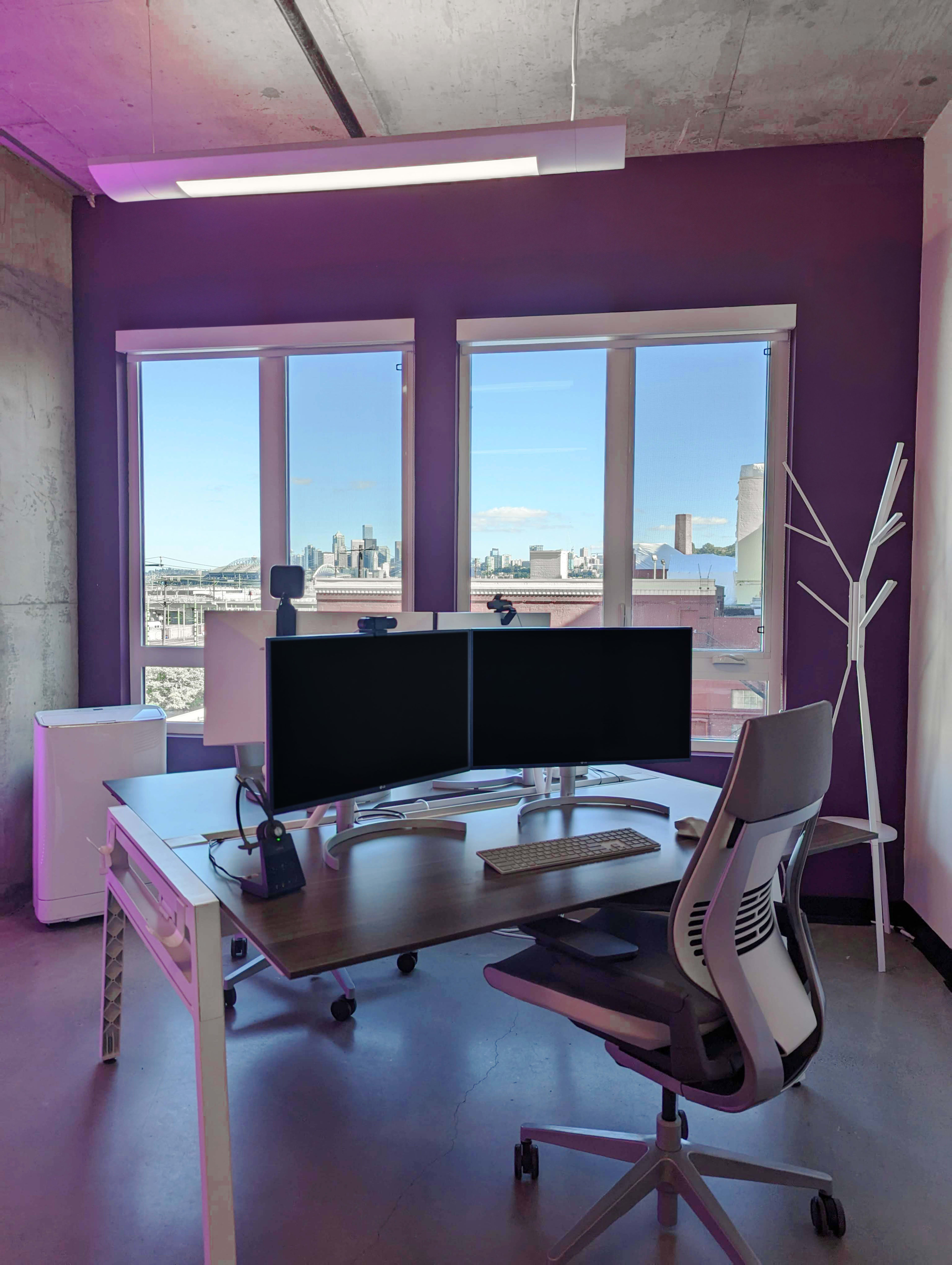 A modern office space features a desk with dual monitors, a keyboard, and a chair, set against a backdrop of large windows revealing a city skyline.