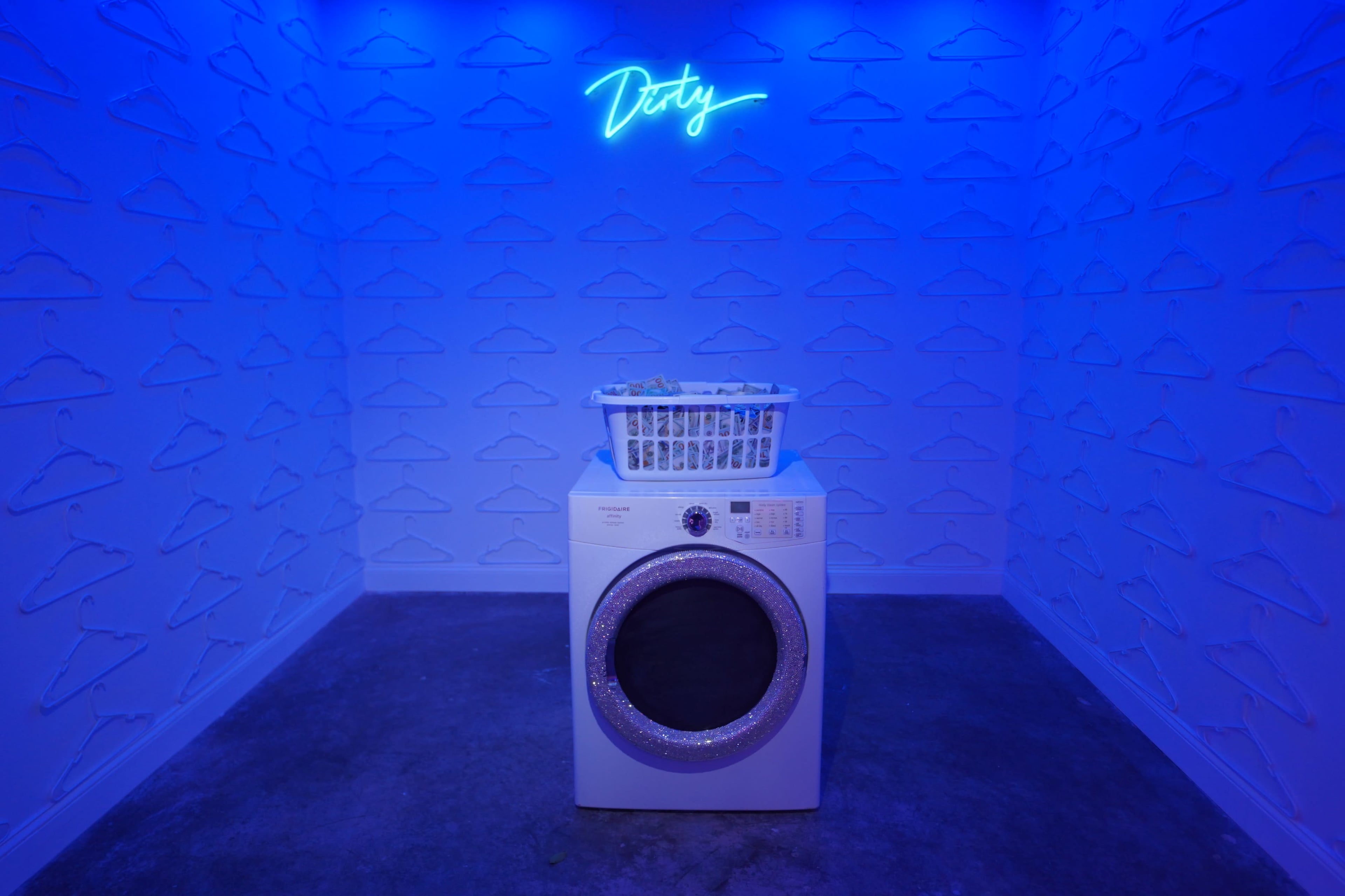 A washing machine with a clothes basket sits against a blue-lit wall adorned with hanger outlines.