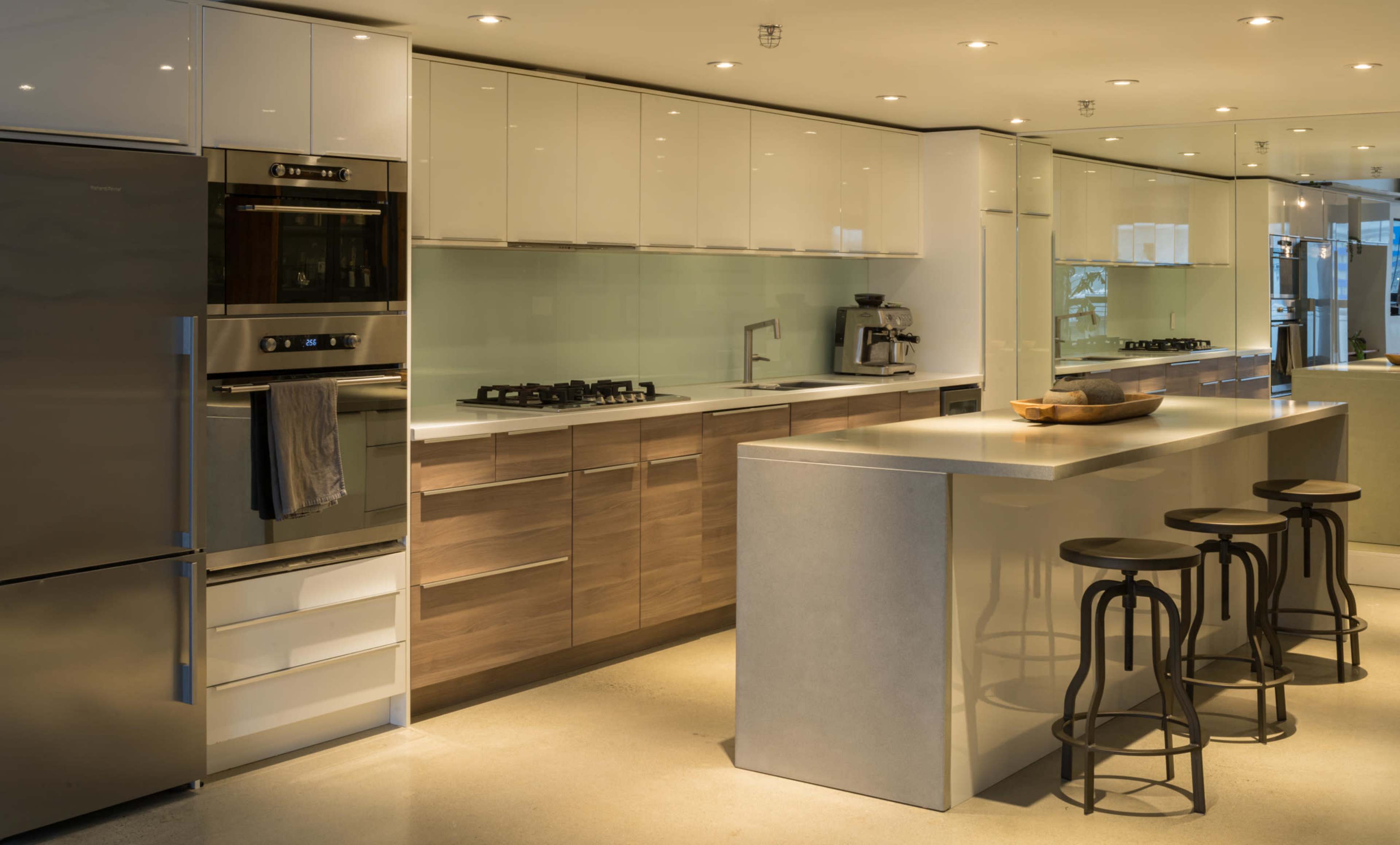 The image shows a modern kitchen with sleek cabinetry, stainless steel appliances, and a large island with stool seating.