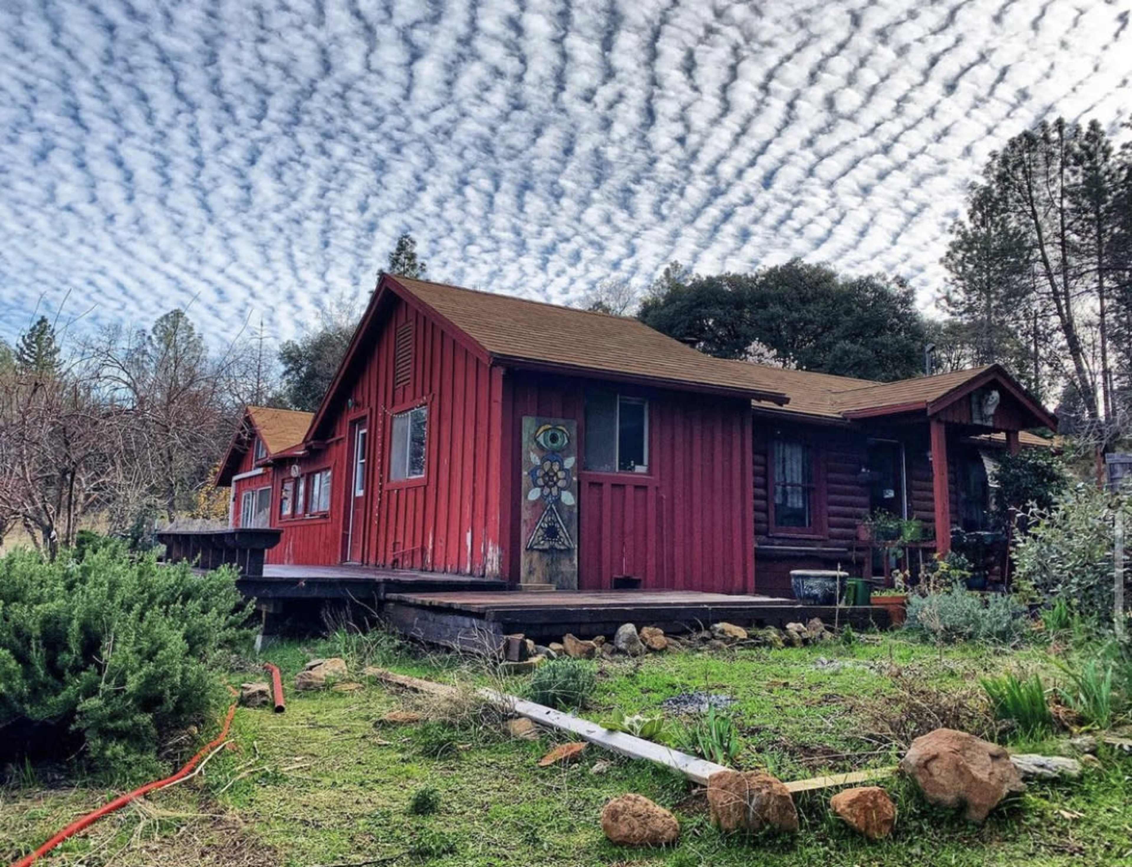 A red wooden cabin with a sloped roof sits on a grassy plot under a patterned cloud-filled sky.