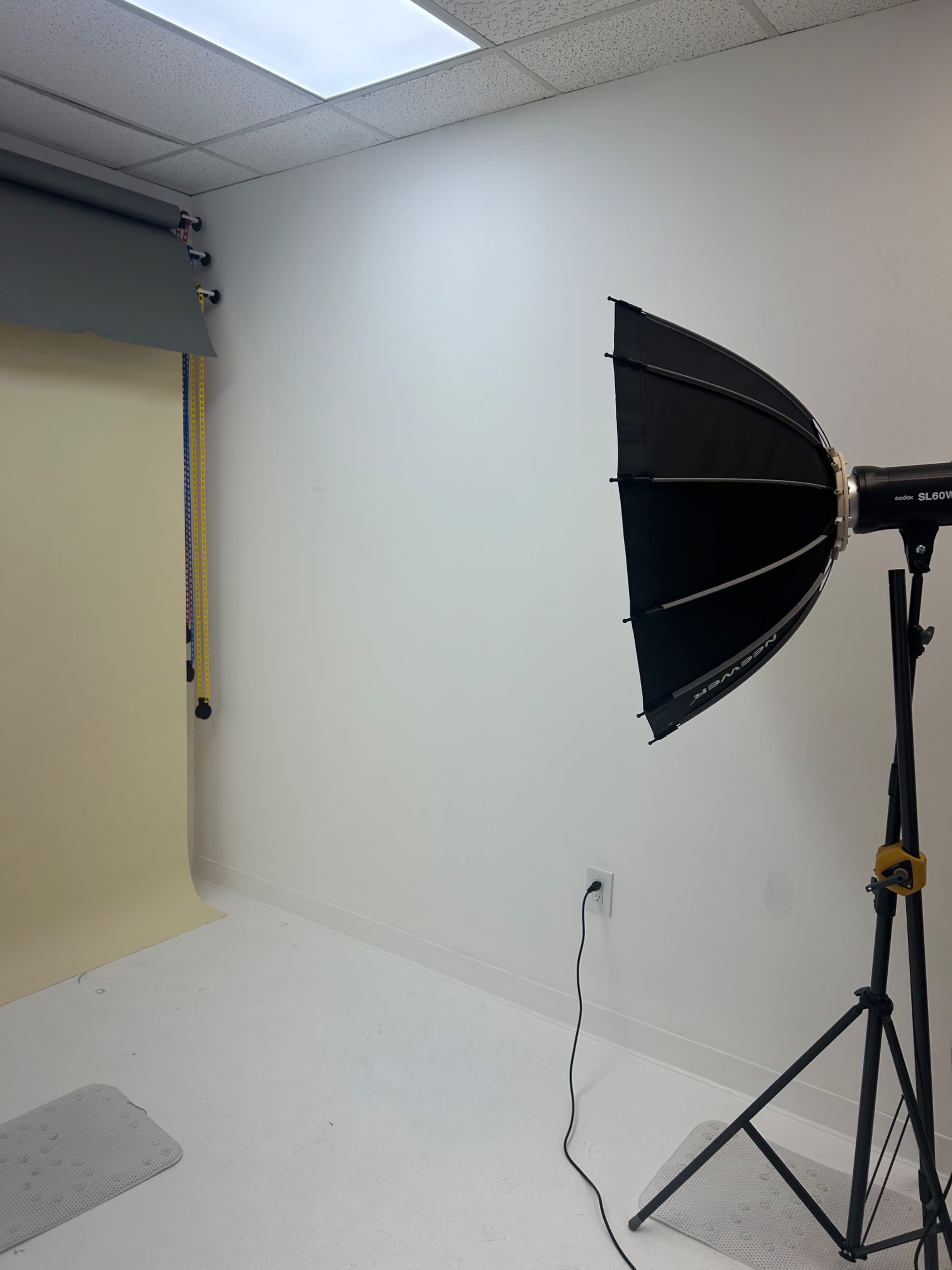 The image shows a photography studio with a light modifier on a stand, a backdrop setup, and plain white walls.