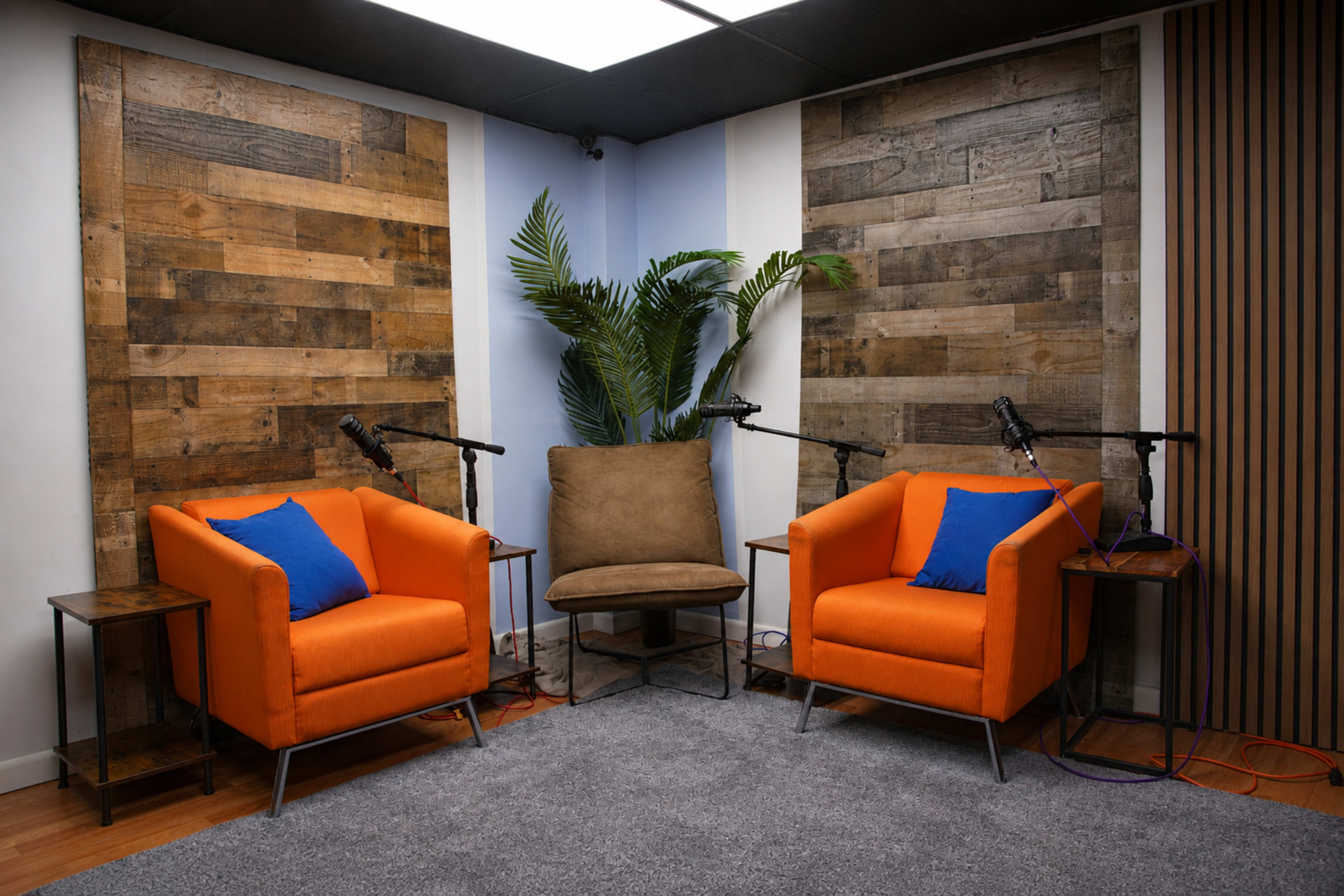 The scene features a small podcast studio with three chairs, two orange armchairs with blue pillows and a brown chair, set against walls made of wooden panels and a backdrop of a palm plant.