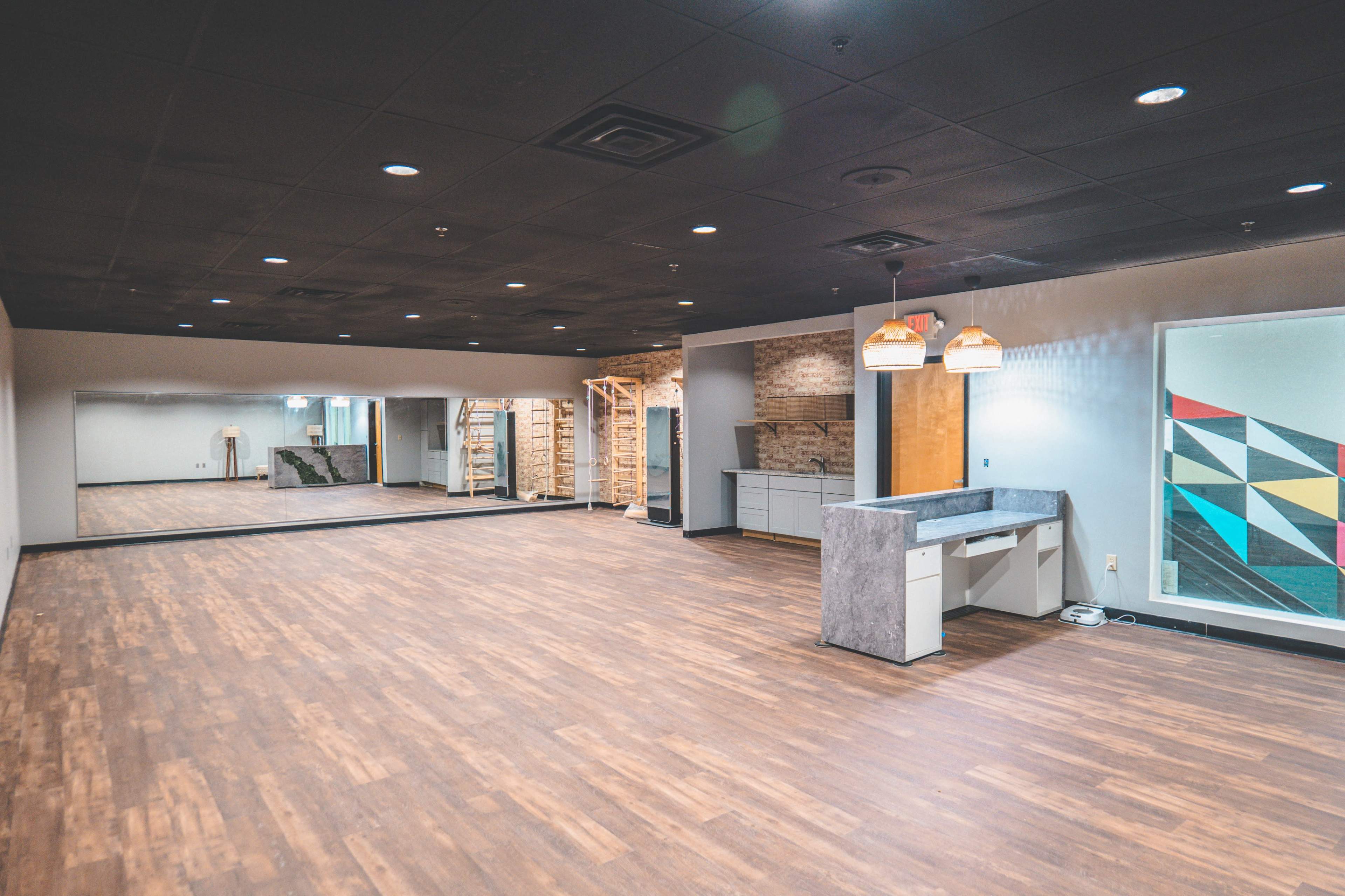 An empty room features wood flooring, a mirrored wall, a small counter area, and modern light fixtures.