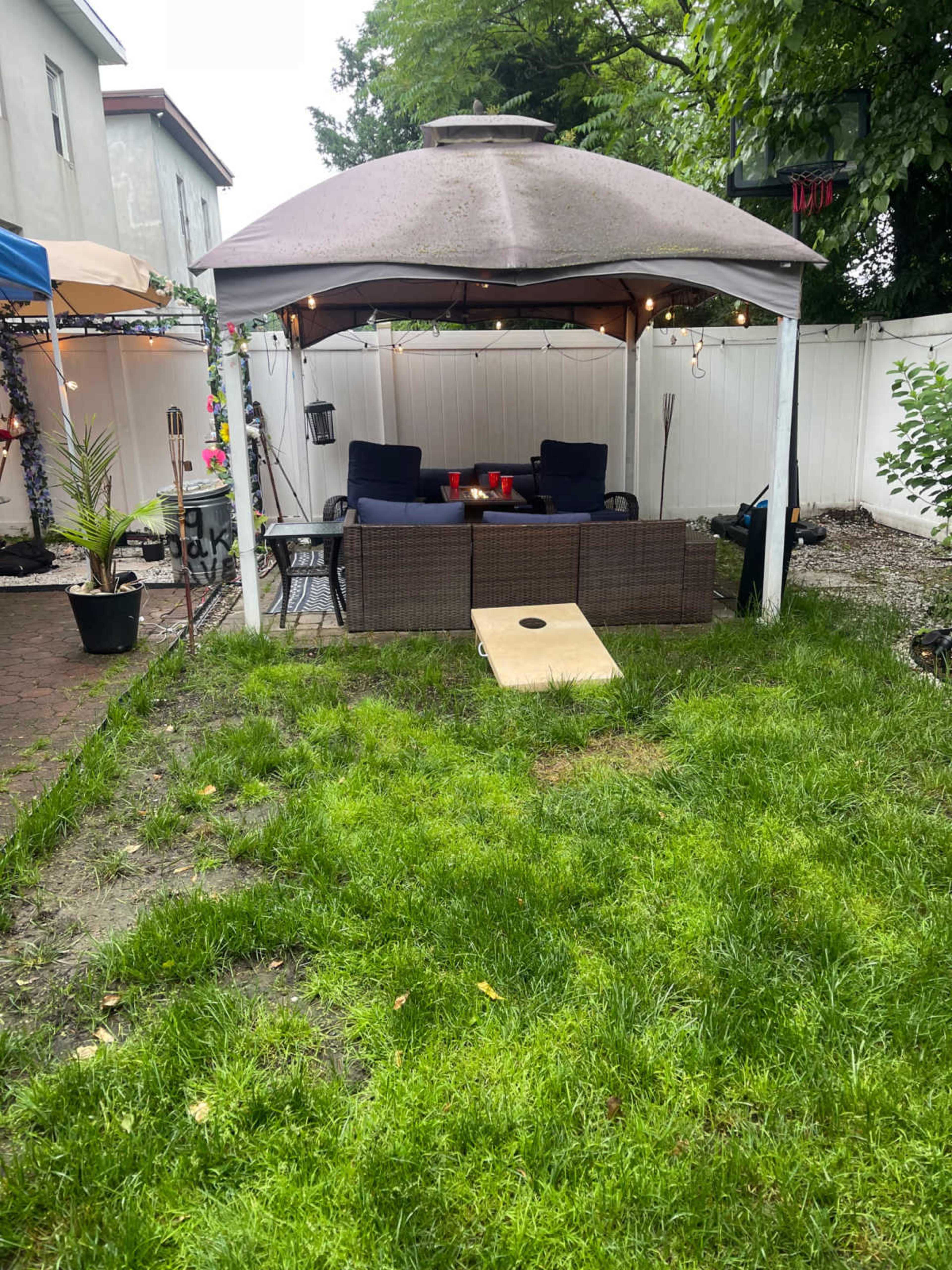 A backyard scene features a gazebo with seating, a cornhole board, and a grassy area surrounded by a white fence.