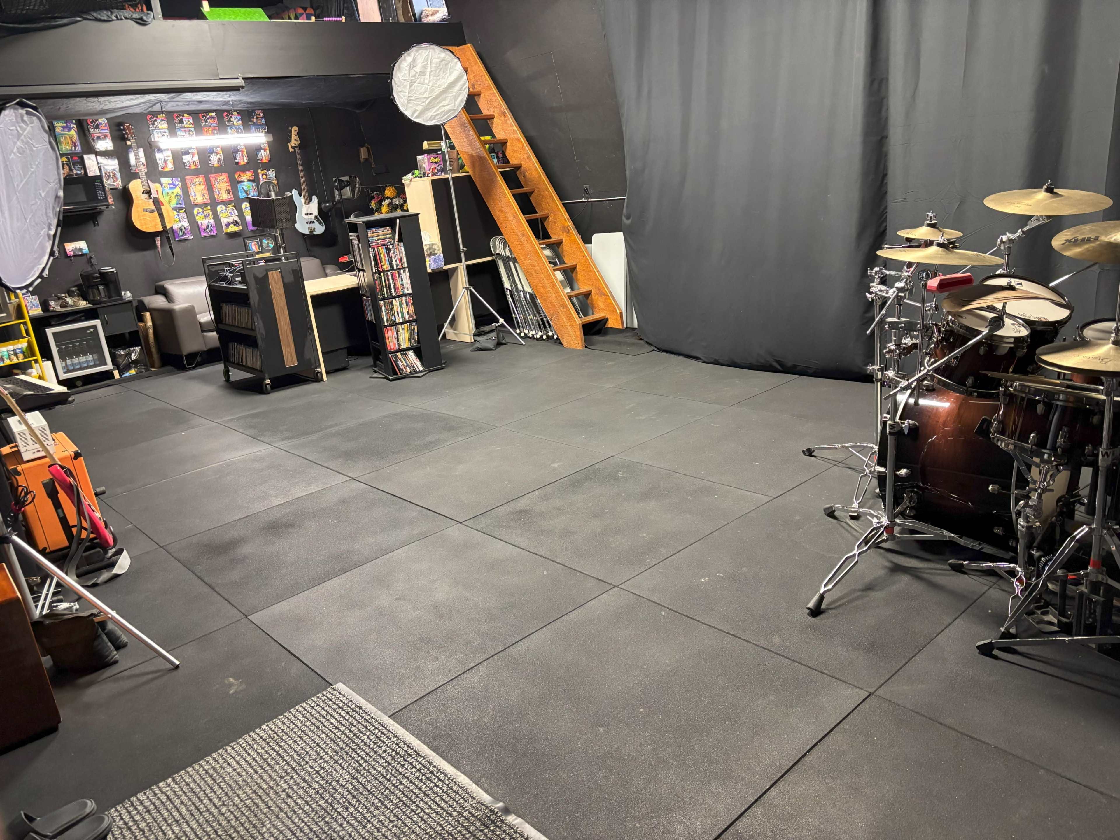 The image shows a music rehearsal space with a drum set on one side, shelving units, and a staircase leading to a higher level, all set against a dark backdrop.