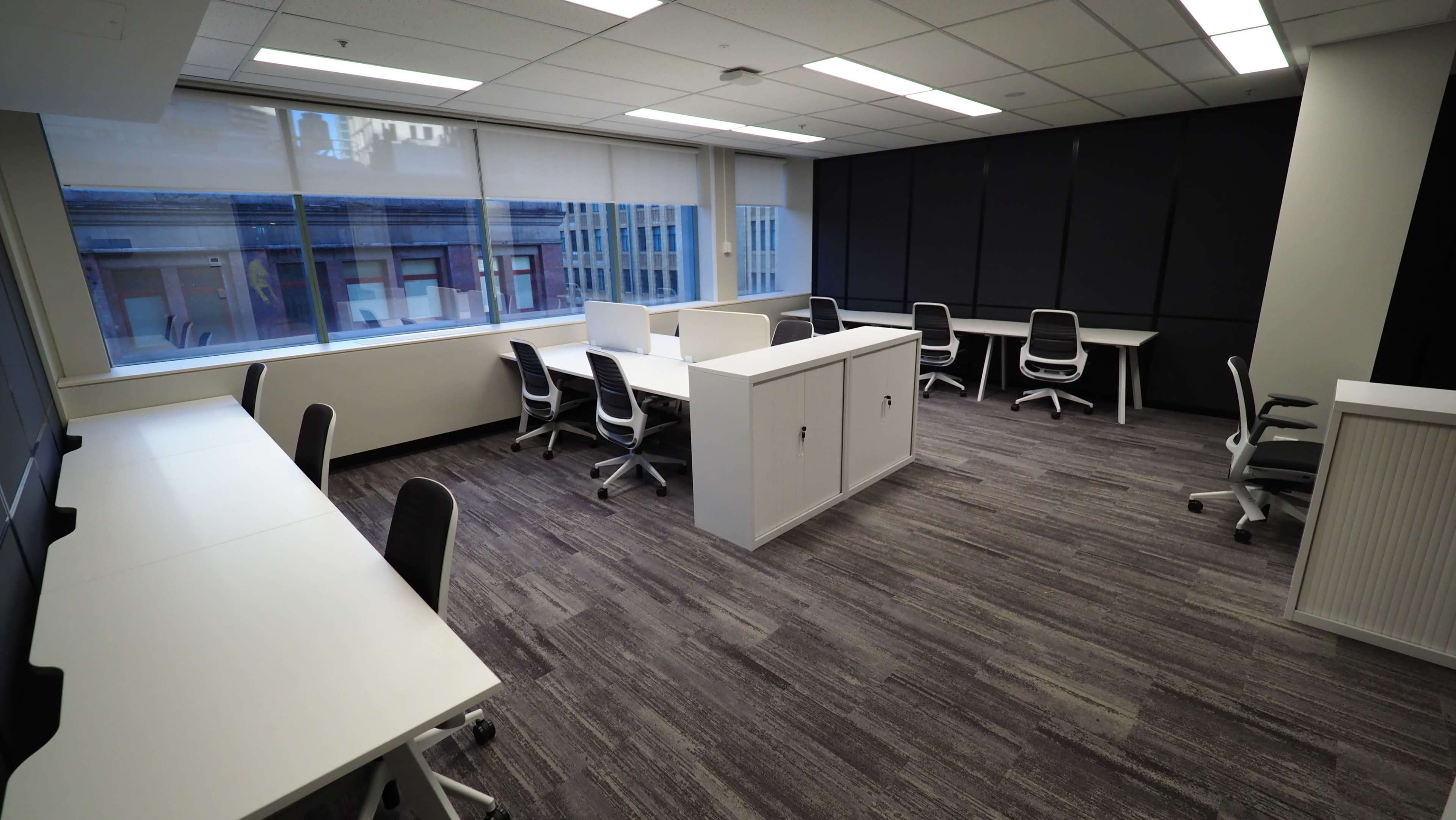 The image shows a modern office space with several white desks and ergonomic chairs arranged near large windows.