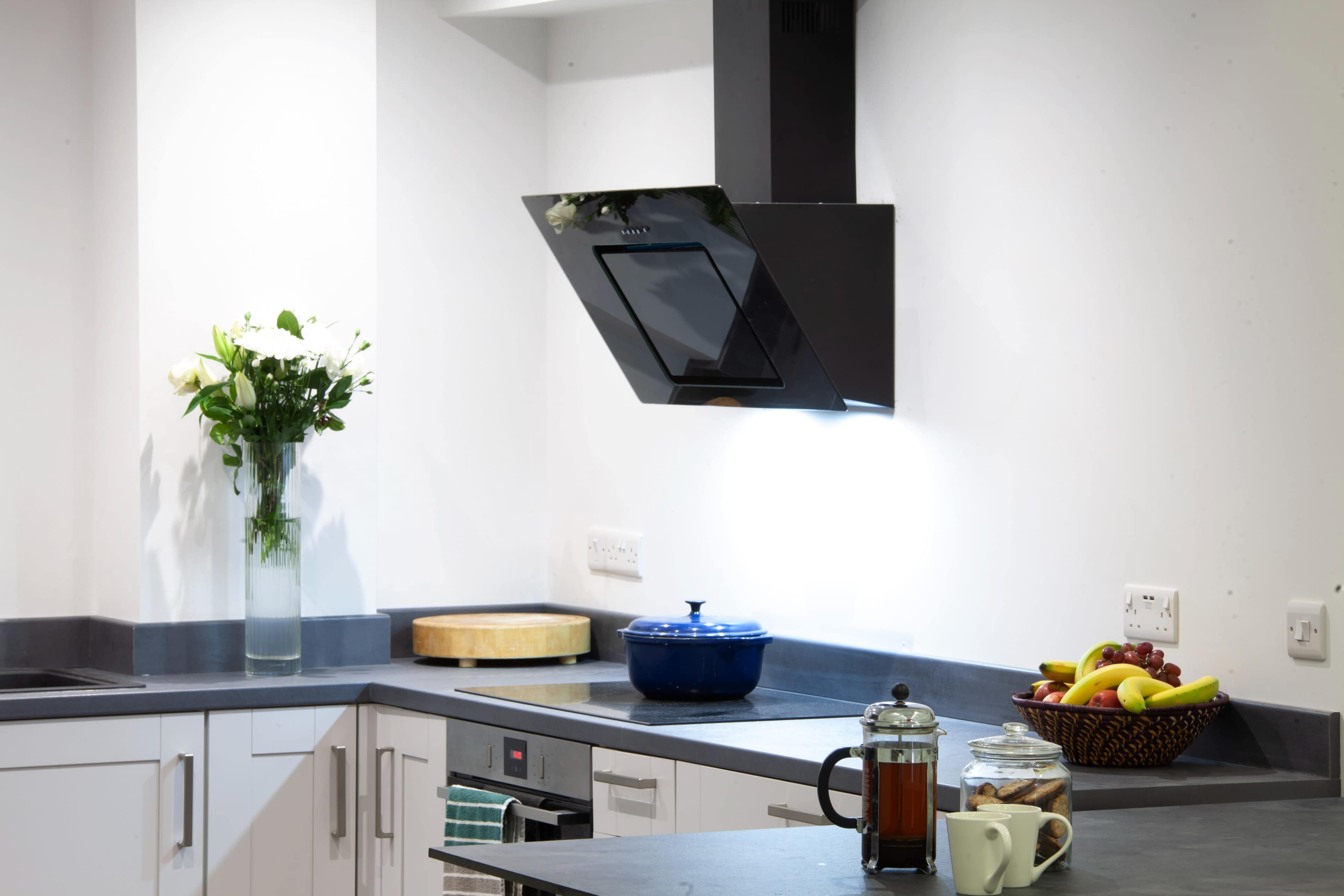 A modern kitchen features a black exhaust hood above a stove, with a blue pot on the countertop and a basket of fruit nearby.