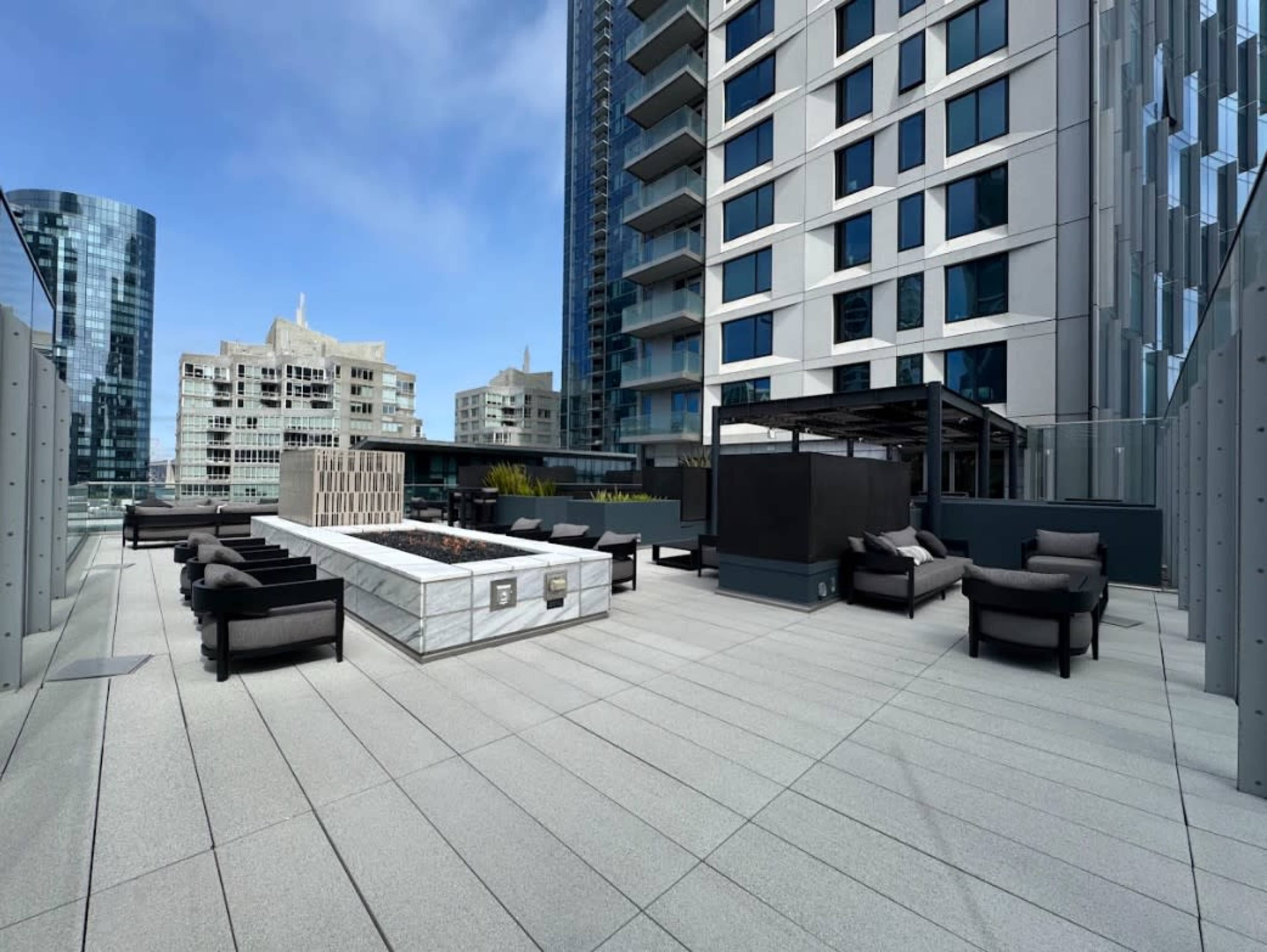 The image shows a modern rooftop terrace featuring seating areas, a fire pit, and views of surrounding buildings.