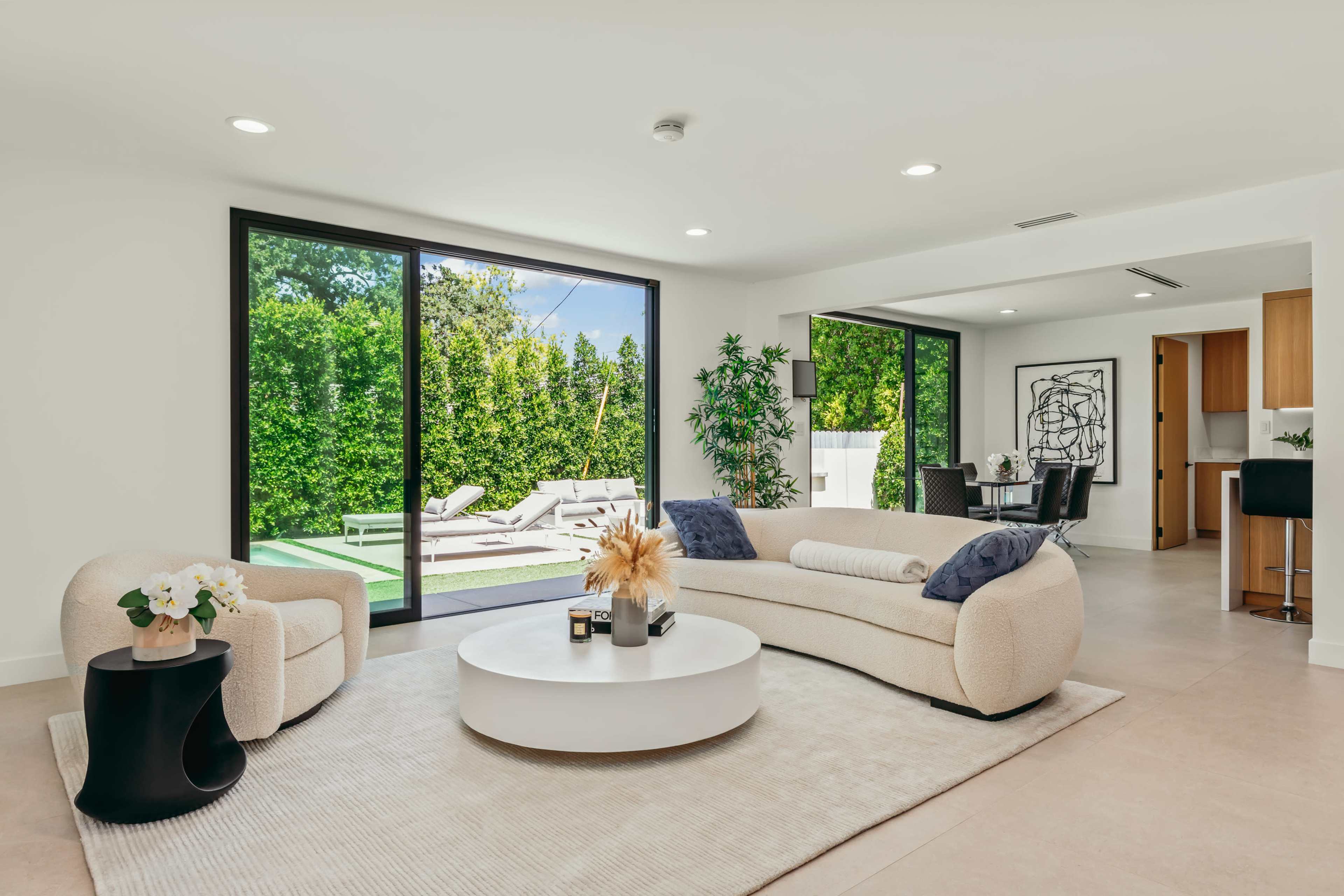 The image features a modern living room with a curved sofa and a round coffee table, facing large glass doors that open to an outdoor patio area.