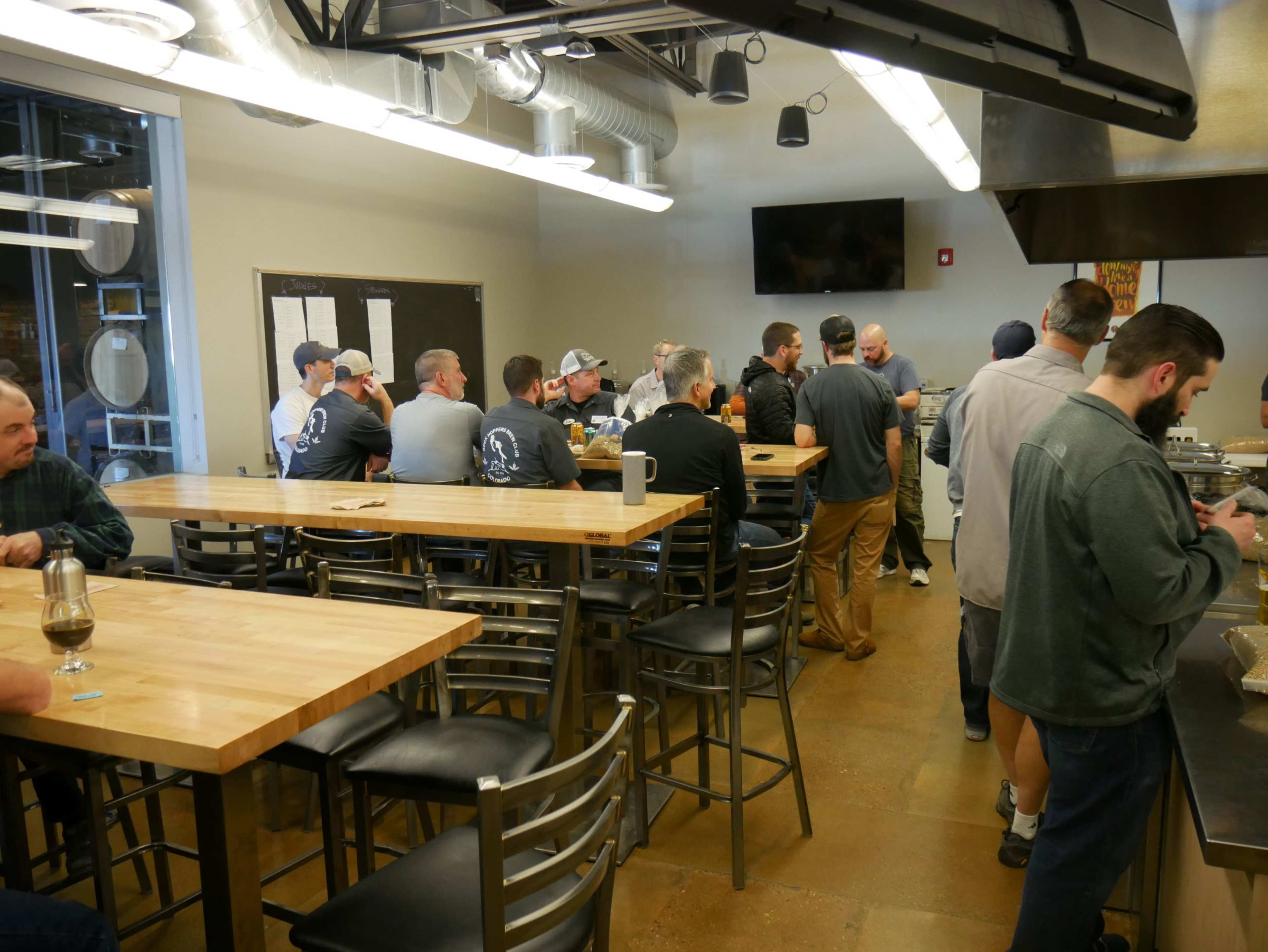 A group of people are gathered in a spacious brewery interior, with several seated at a long wooden bar and others standing near a counter.