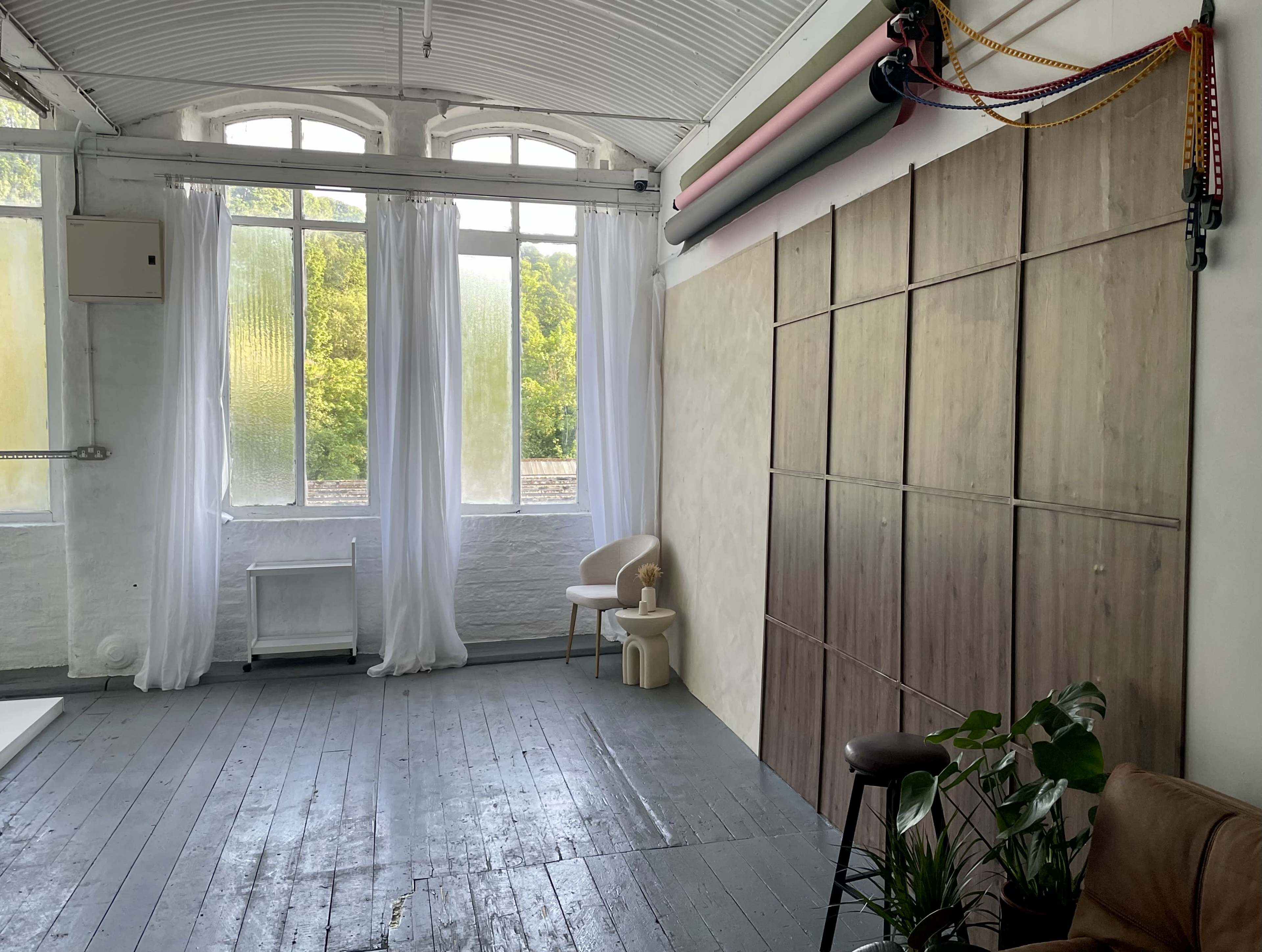 The image shows a spacious studio with large windows, sheer white curtains, and wooden flooring, featuring a beige wall and a small chair beside a stool.