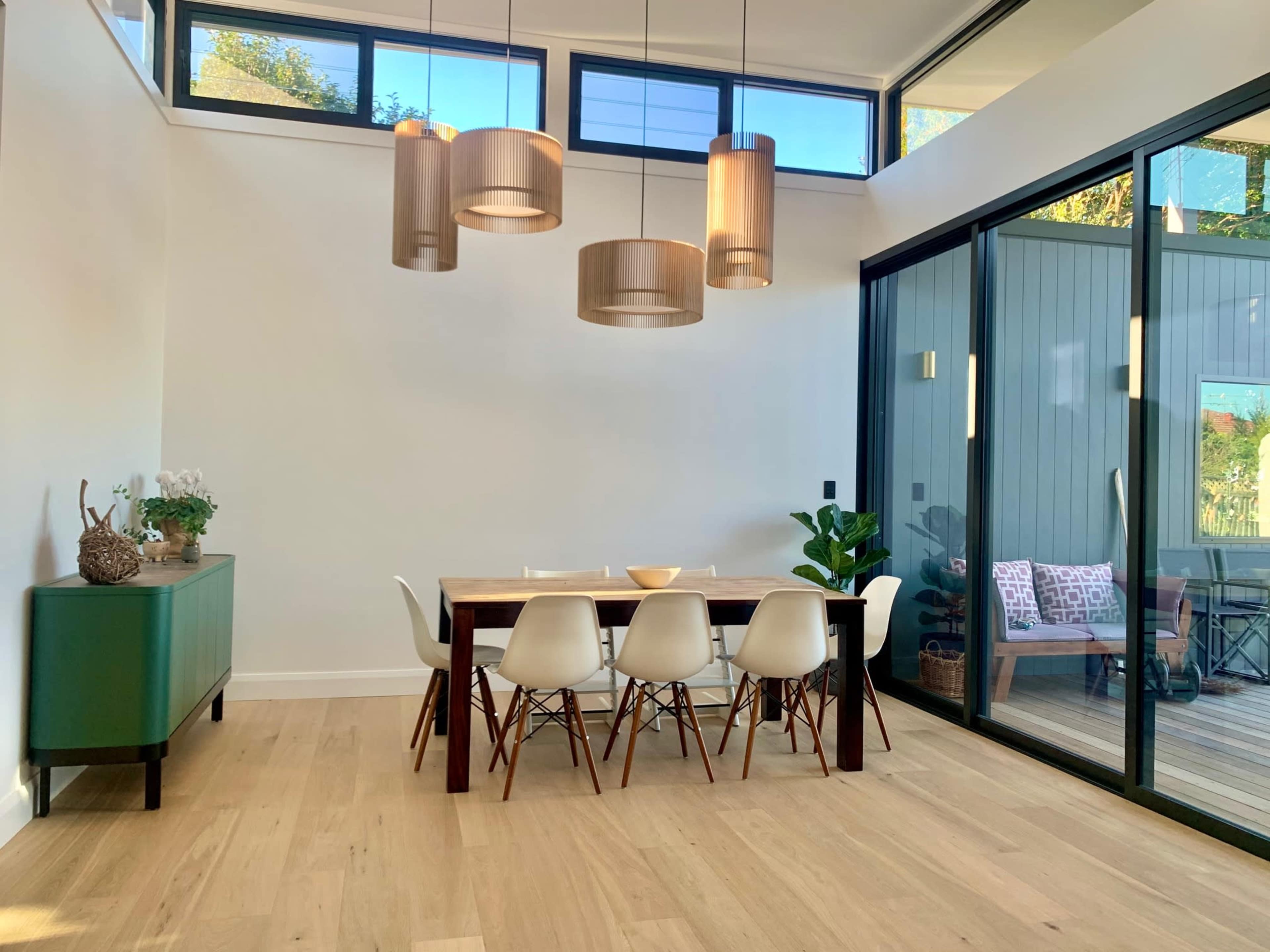 A modern dining area features a wooden table surrounded by white chairs, with pendant lights overhead and large glass doors opening to an outdoor space.