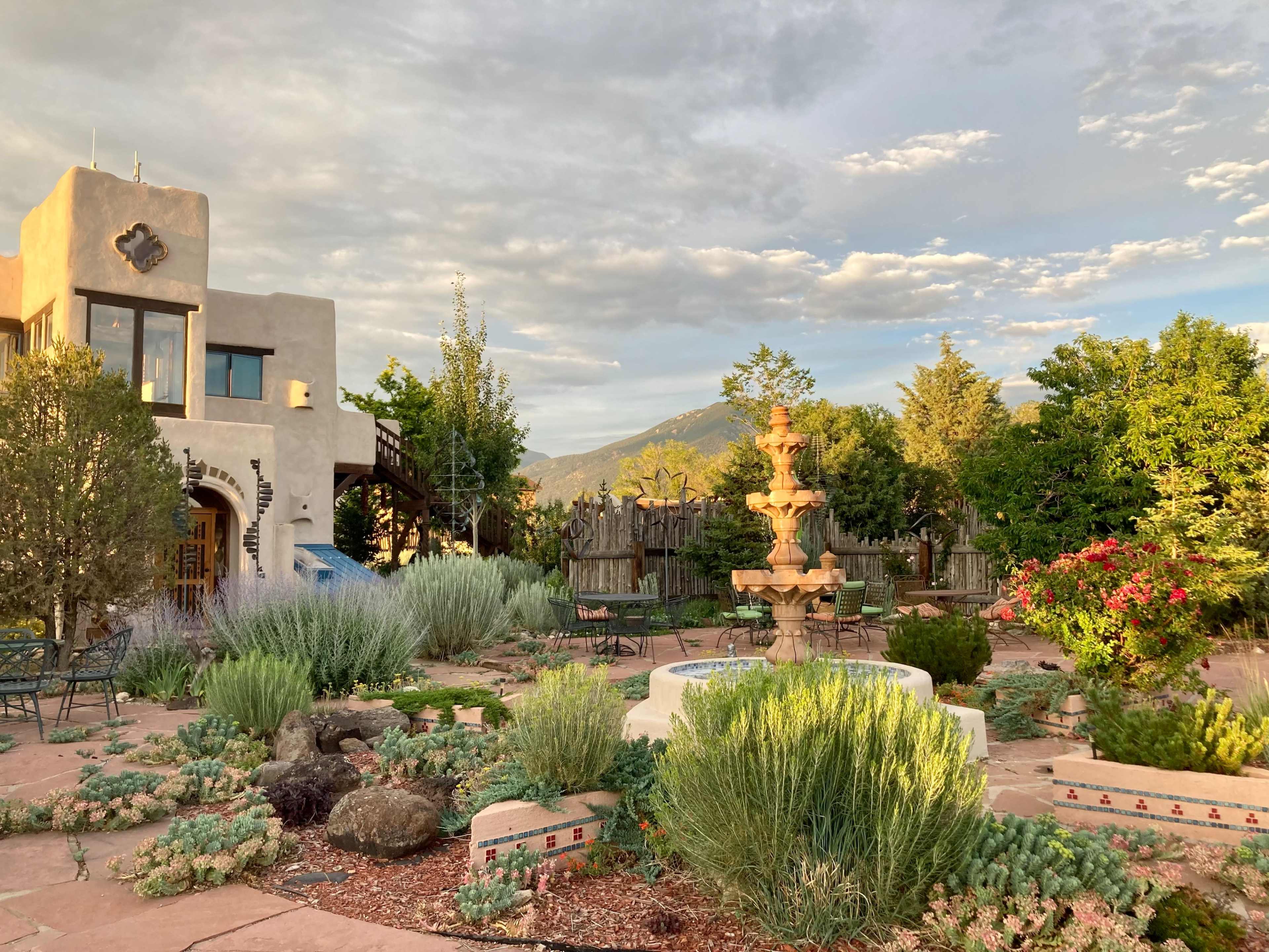 A landscaped garden features a multi-tiered fountain surrounded by various shrubs and plants, with a backdrop of mountains and an adobe-style building.