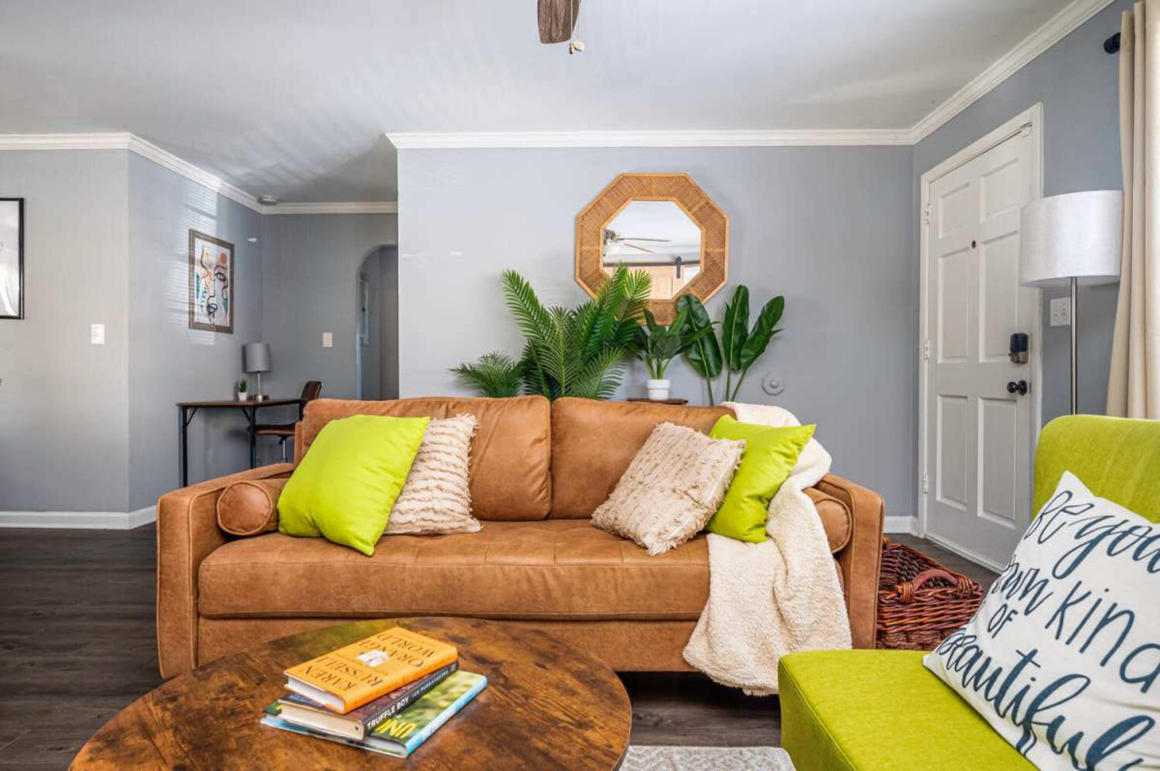 A living room features a brown sofa with bright green pillows, a round wooden coffee table topped with books, and a corner with potted plants and a lamp.