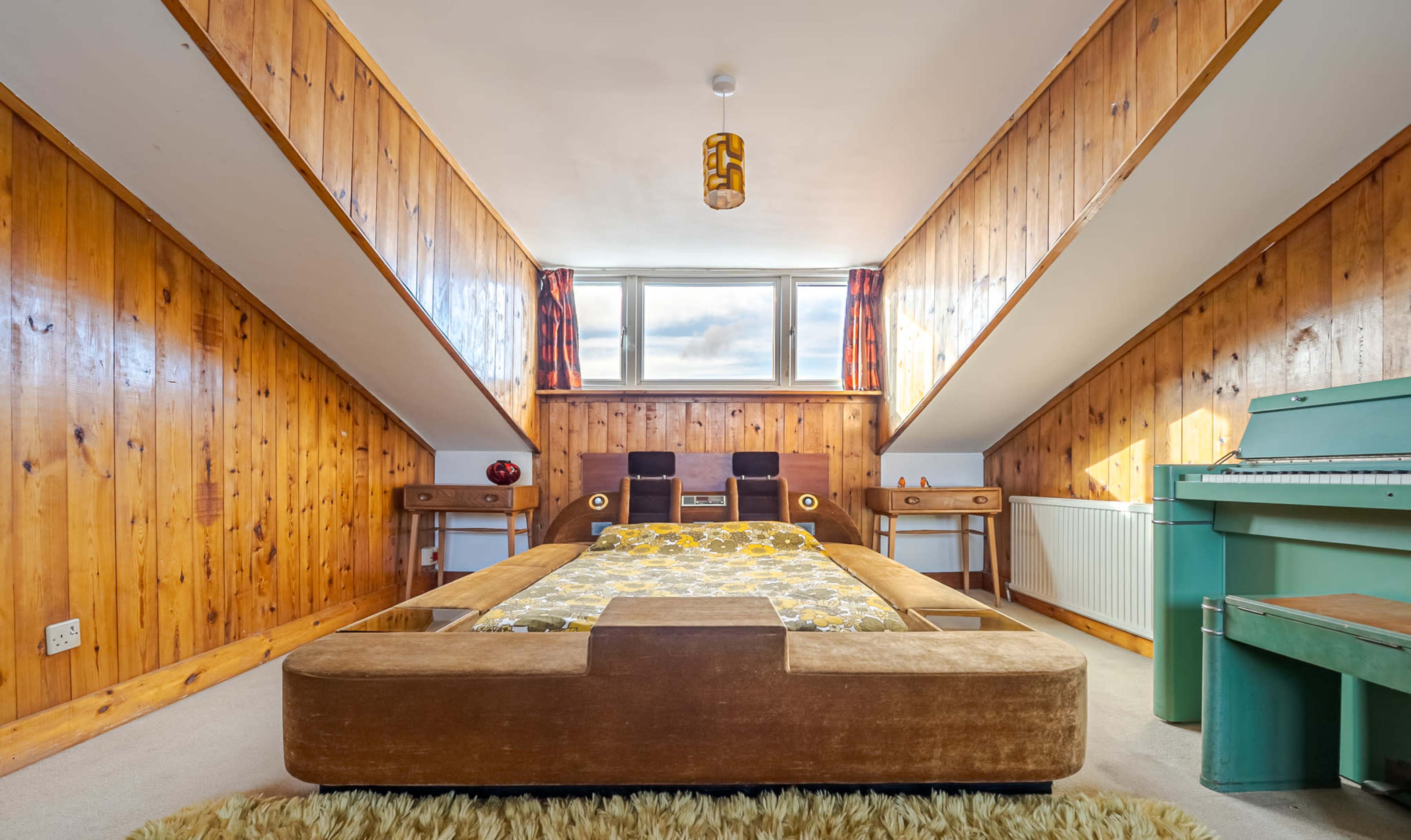 The image shows a bedroom with wooden paneled walls, a low bed in the center, two chairs, a small table, and a piano near a window.