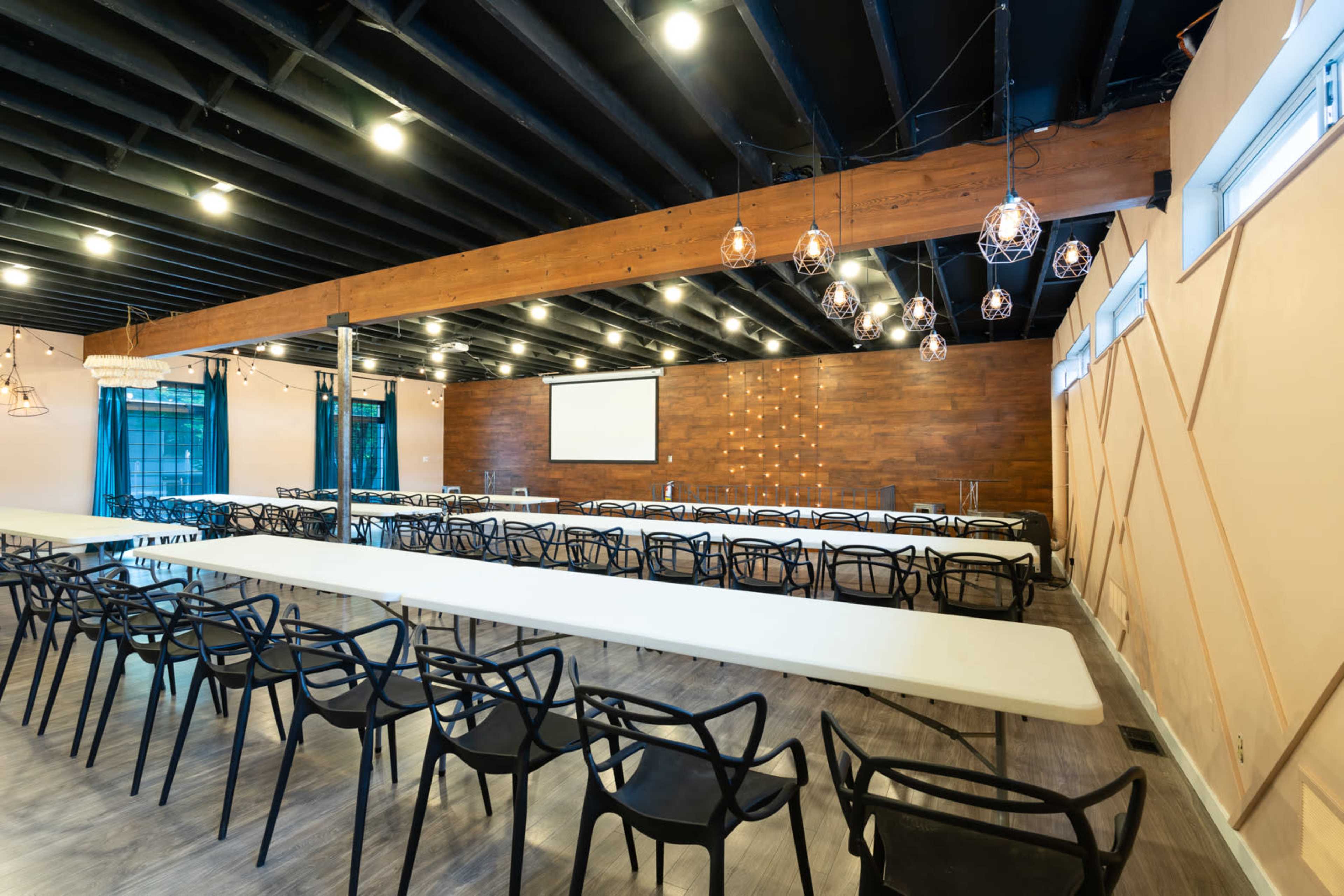 A large, empty conference room with white tables arranged in rows, black chairs, and a wooden accent wall featuring a projector screen.