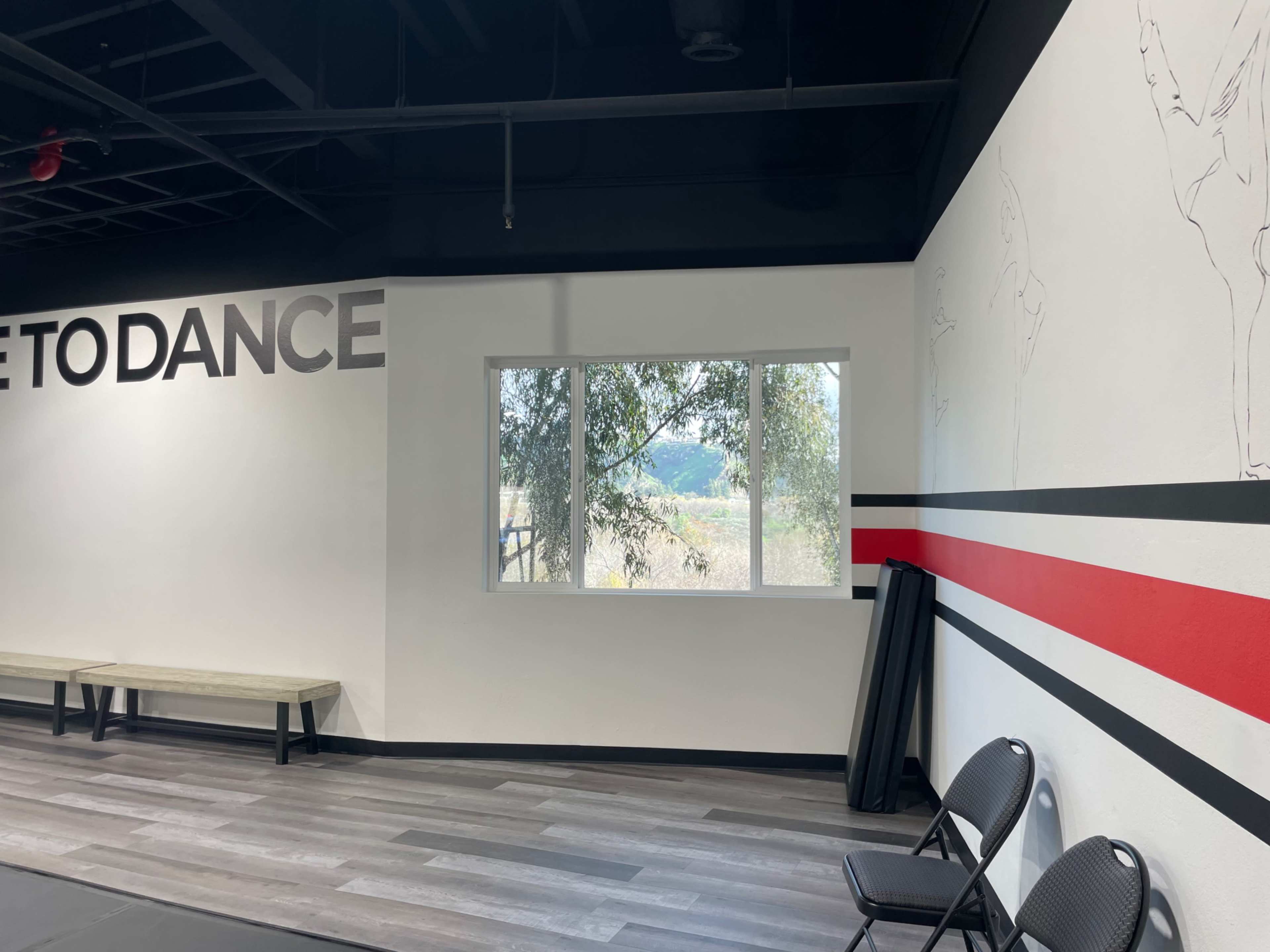 A dance studio interior with a large window overlooking a field, featuring the words "LET'S DANCE" on the wall and a simple seating area.