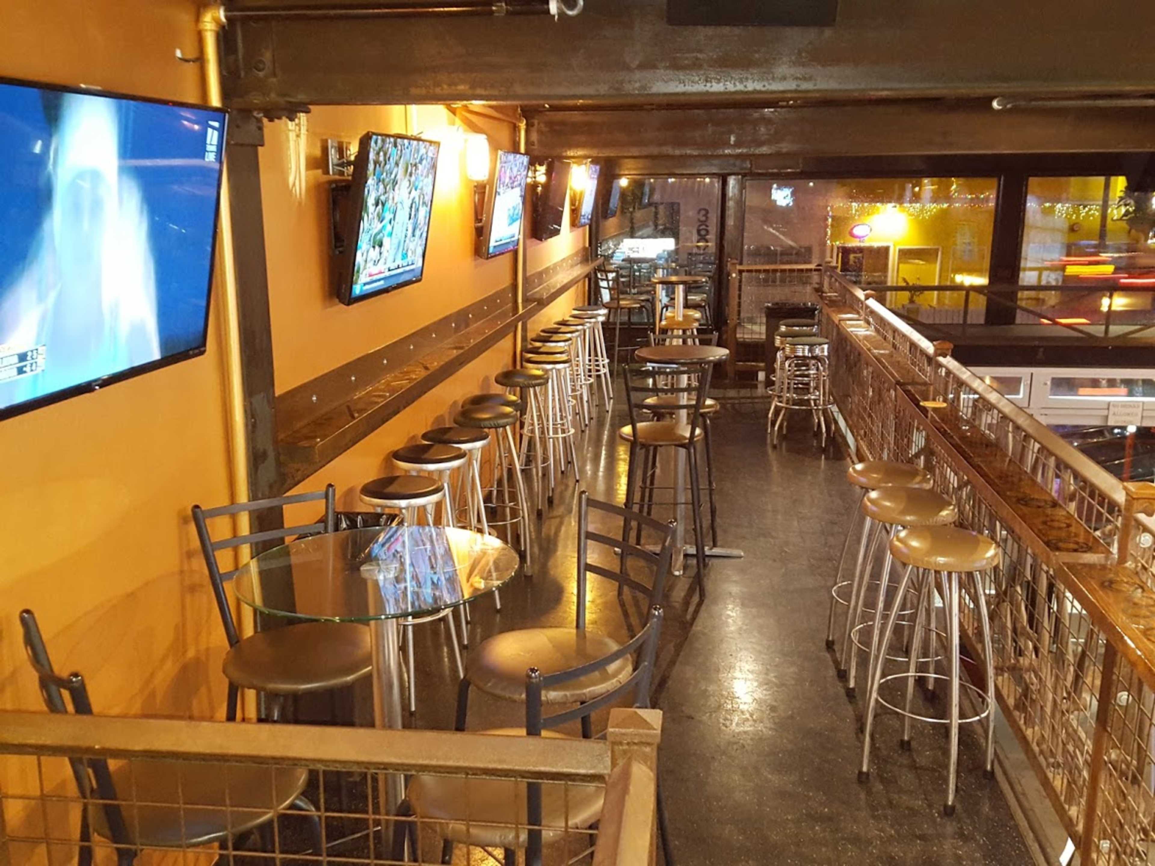 The image shows a narrow bar area with high chairs and tables, featuring multiple television screens mounted on the walls.