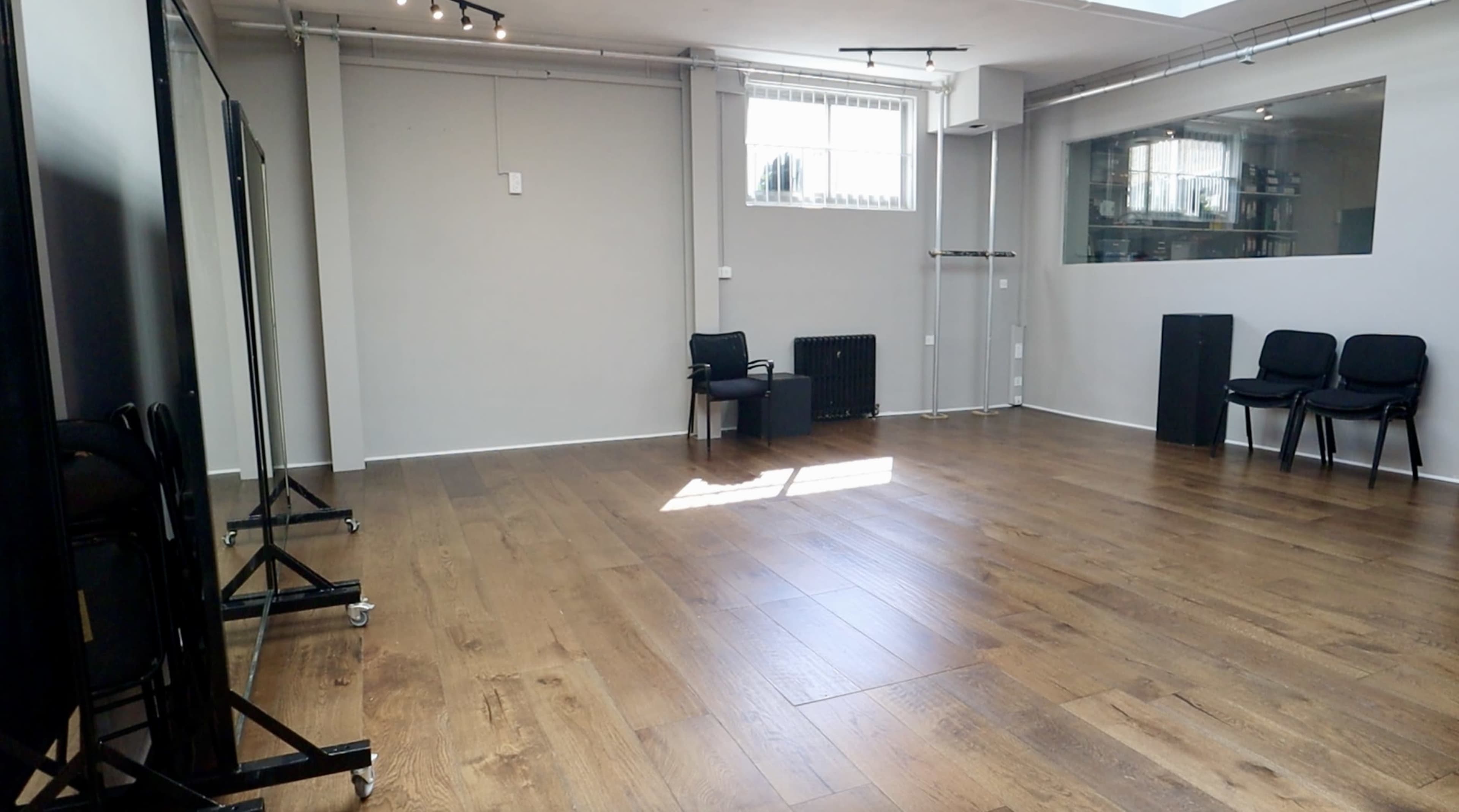 An empty, well-lit room with wooden flooring, a large window, and a few black chairs arranged in the space.