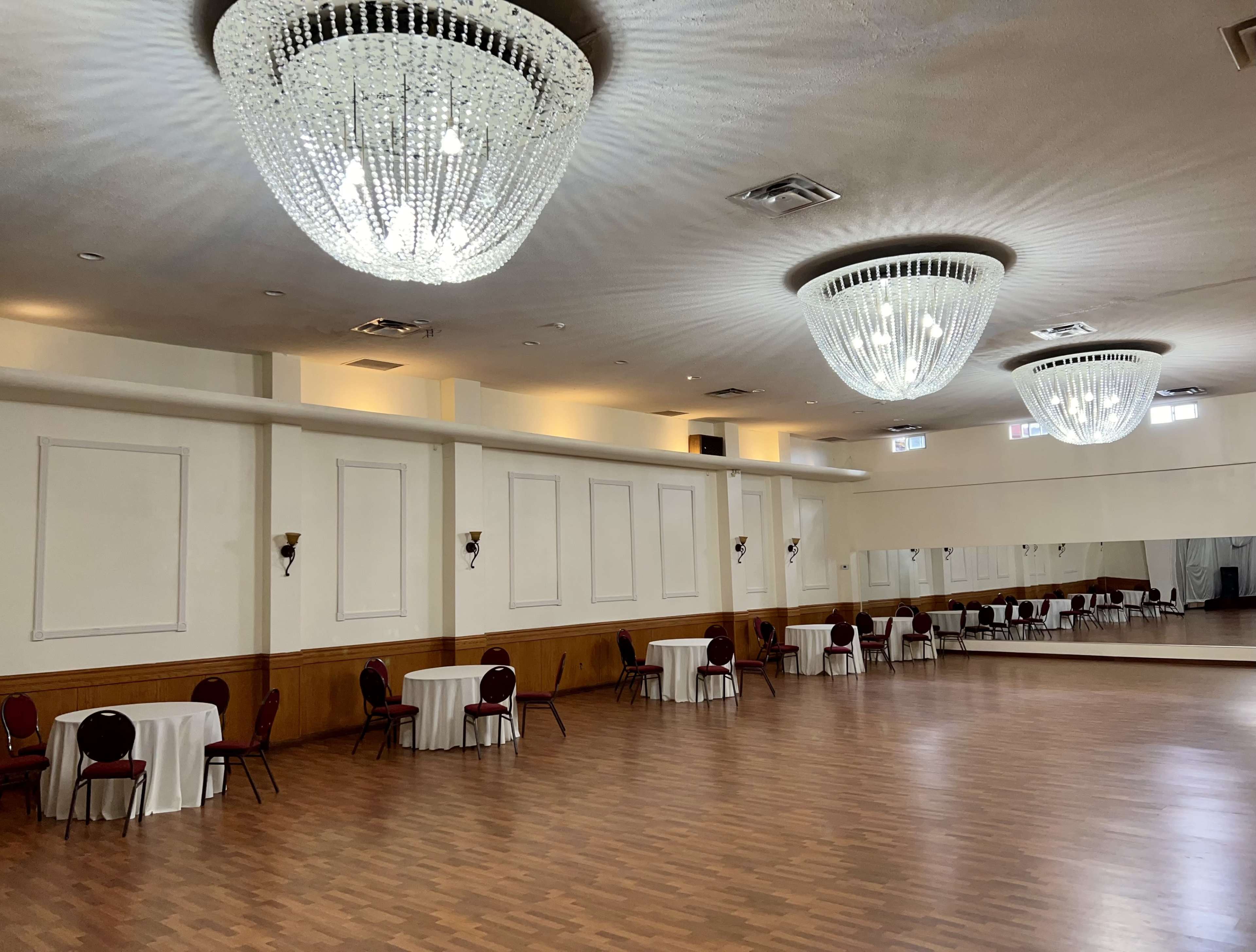 A large, empty banquet hall with two chandeliers, round tables draped in white cloth, and a mirrored wall.