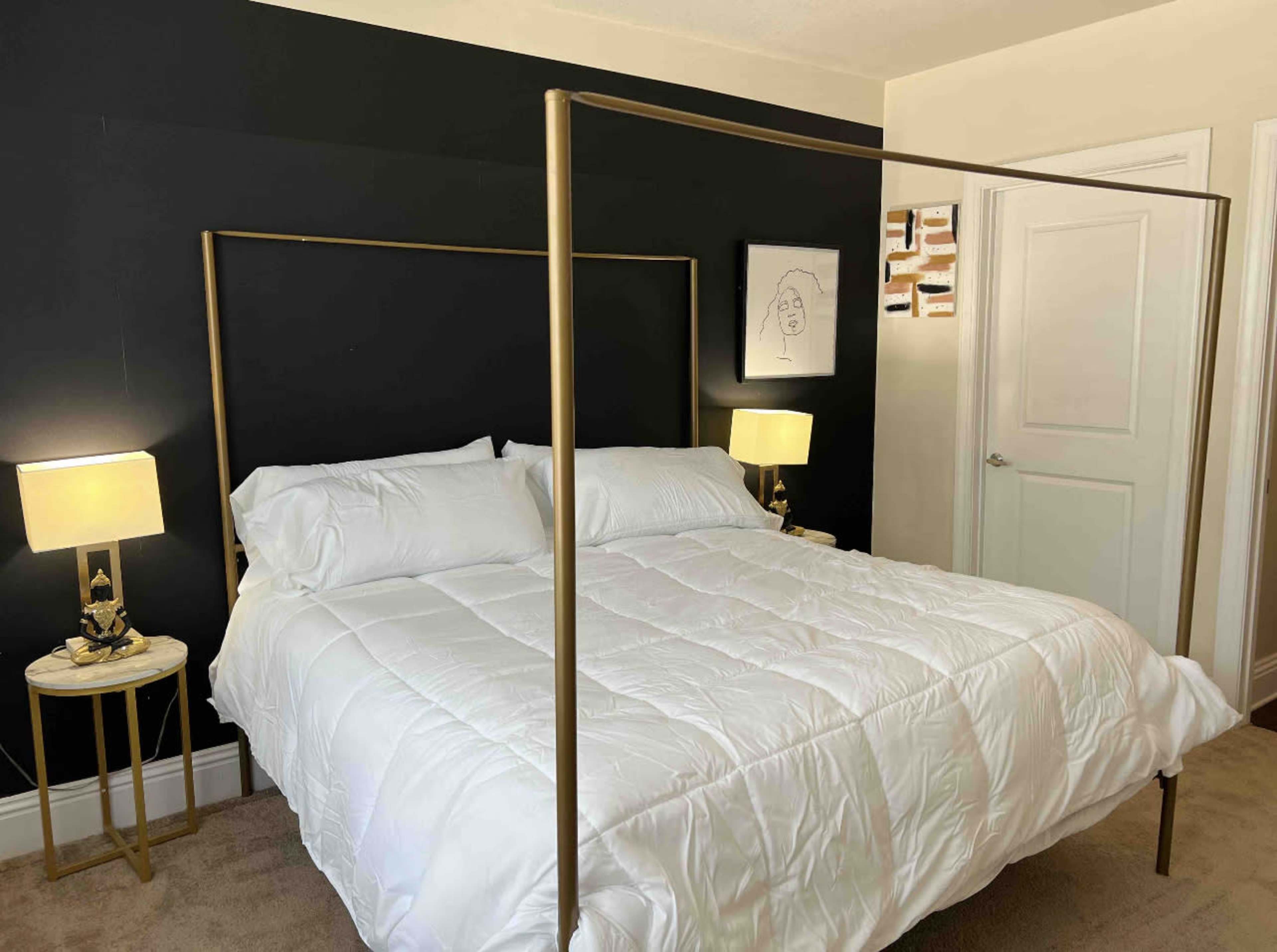 The image shows a neatly arranged bedroom with a four-poster bed, two bedside lamps, and a decorative wall art piece.