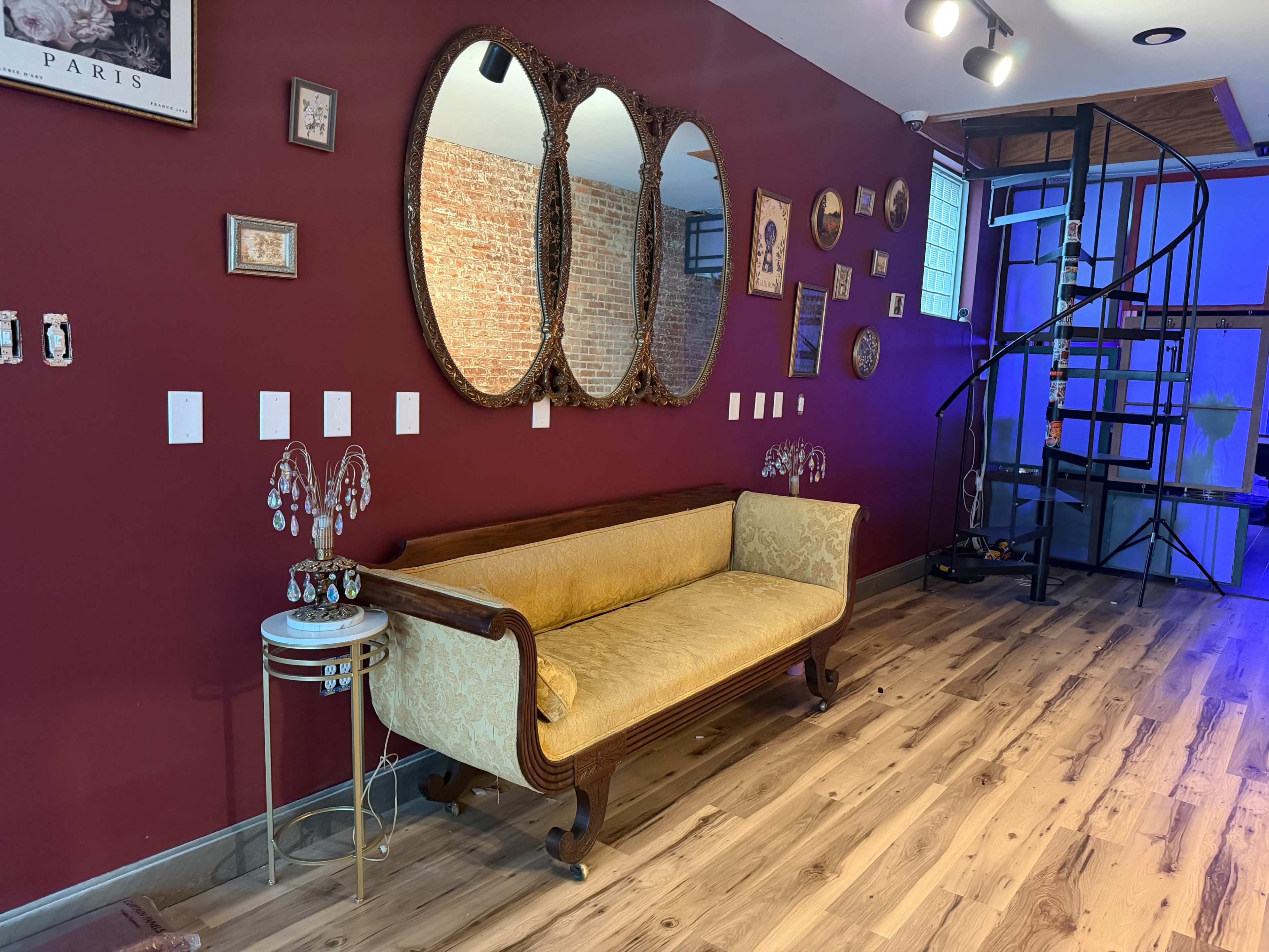 The image features a vintage-style yellow sofa against a maroon wall adorned with mirrors and framed art, alongside a spiral staircase in the background.