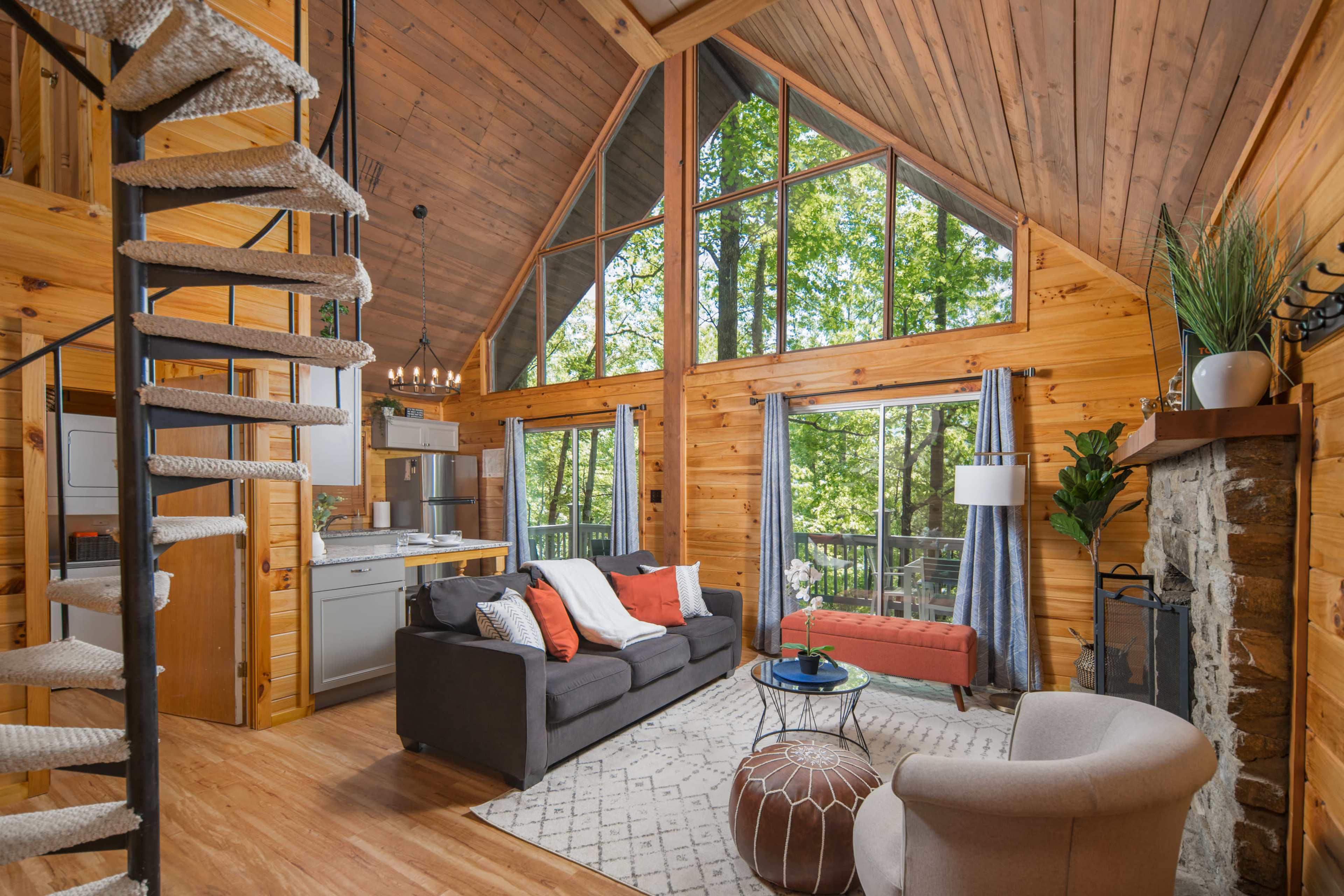 A wooden interior of a cabin with high ceilings, a spiral staircase, a living area featuring a gray sofa, a coffee table, and large windows overlooking greenery.