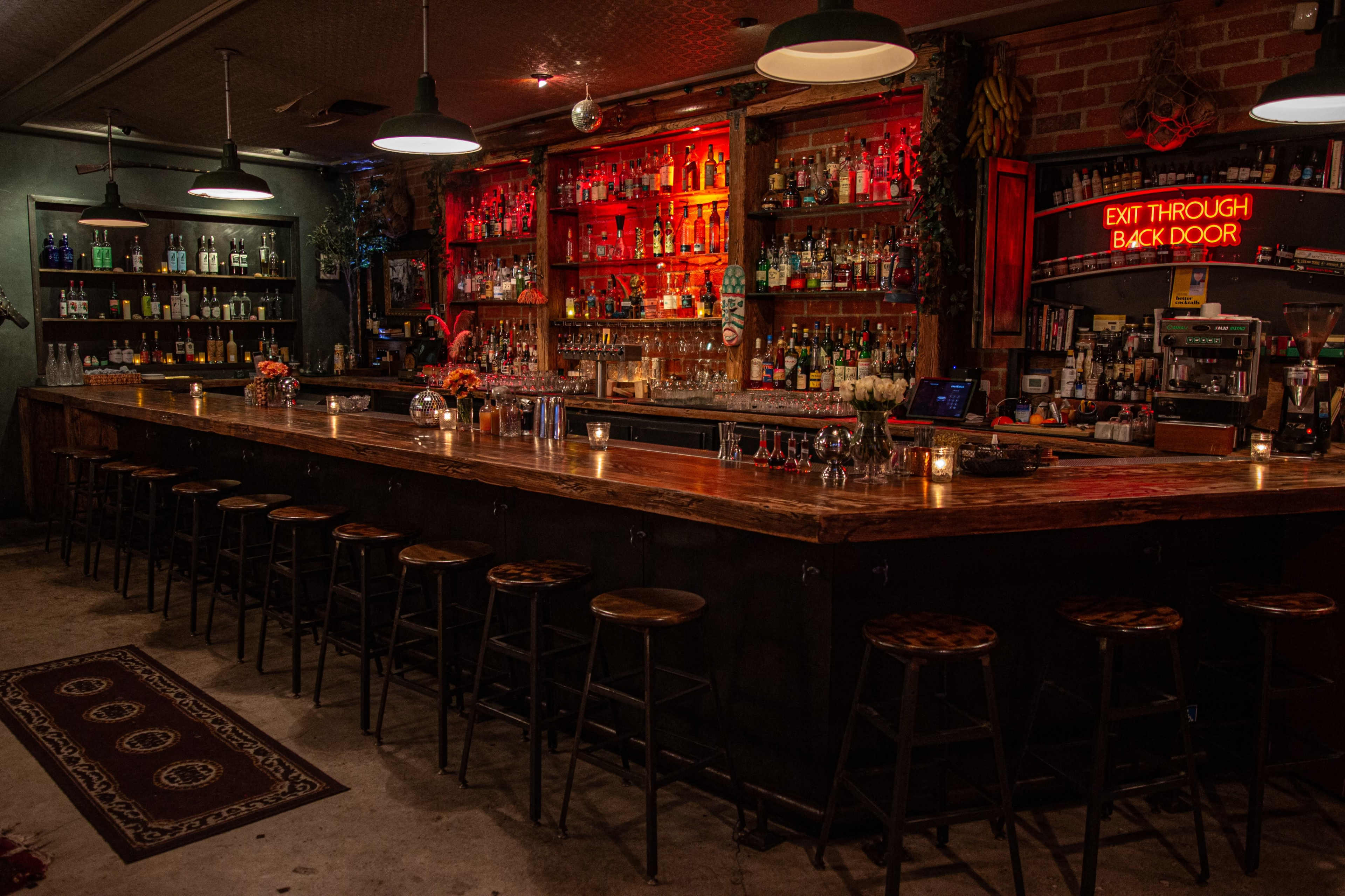 A dimly lit bar features a long wooden counter with several stools, shelves lined with various bottles, and a neon sign that reads "EXIT THROUGH BACK DOOR."