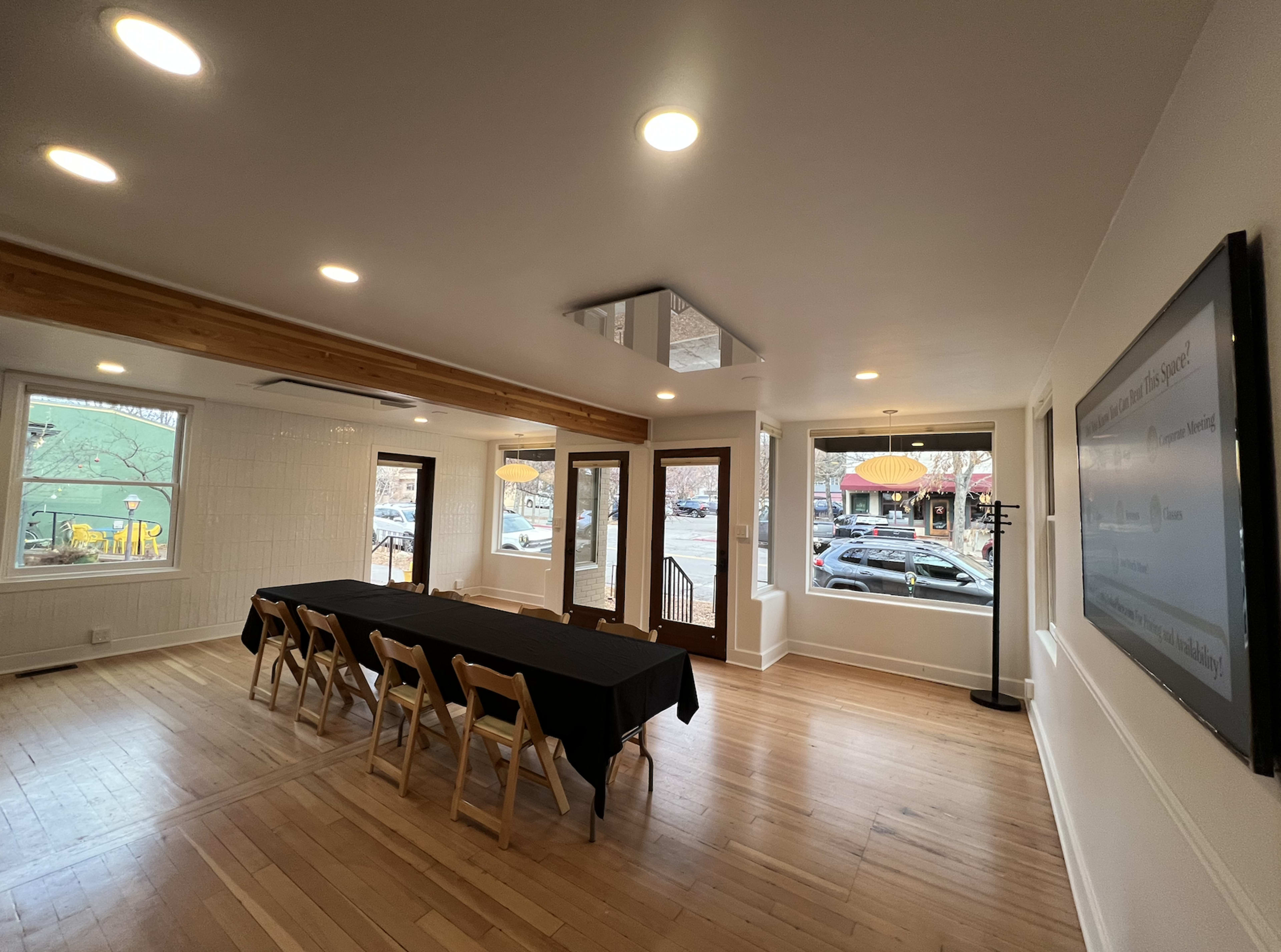 The image shows a well-lit interior of a room featuring a long black table with chairs, large windows, and an inviting entrance.