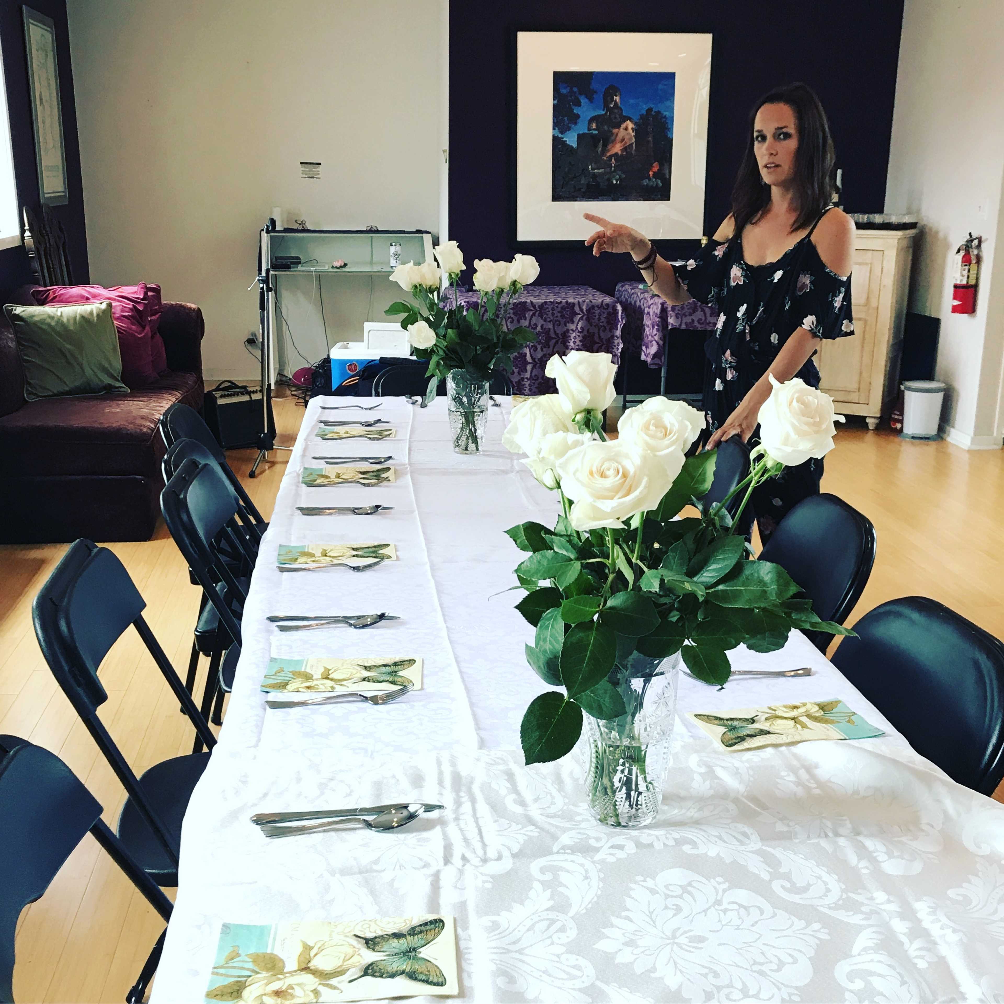 A woman prepares a long dining table adorned with white roses in vases and neatly arranged place settings.